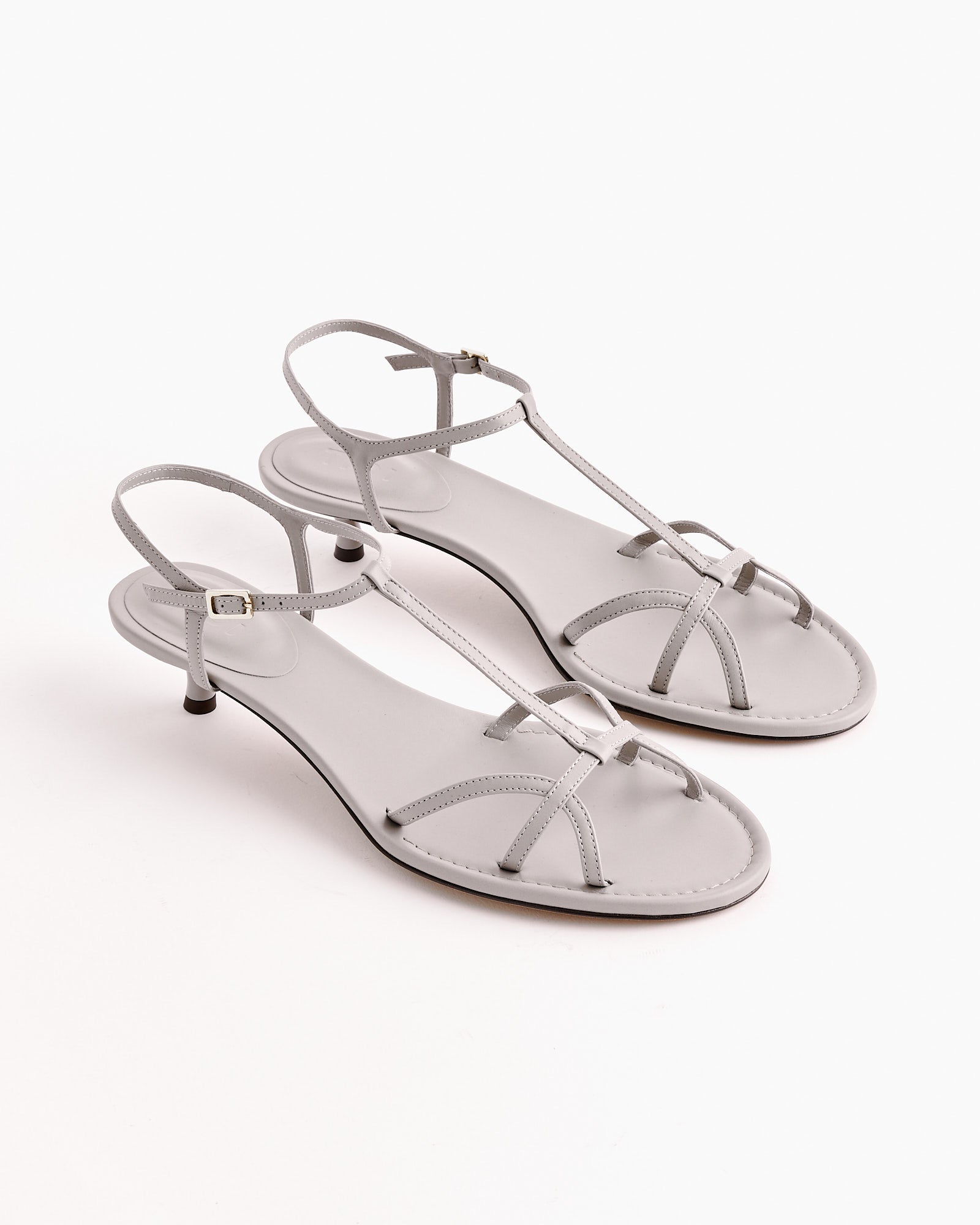 A pair of Tibi Christopher Heel sandals in grey with thin straps, kitten heels, and adjustable ankle buckles in supple calfskin leather, shown on a white background.