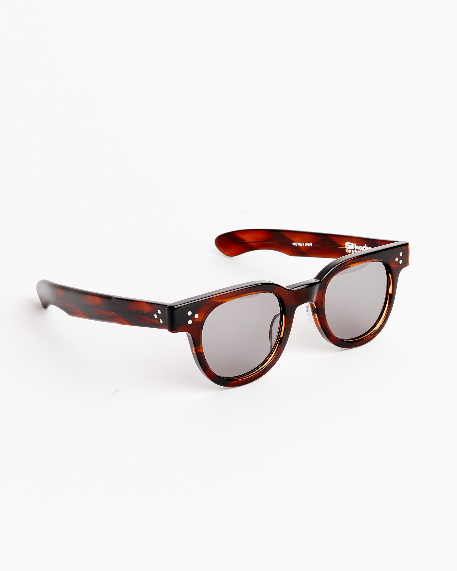 The Frankie Sunglasses in Demi Amber/Grey by Shady Character feature round, dark grey lenses and thick brown tortoiseshell frames with silver accents. Displayed on a white background—contact online chat for sizing help.