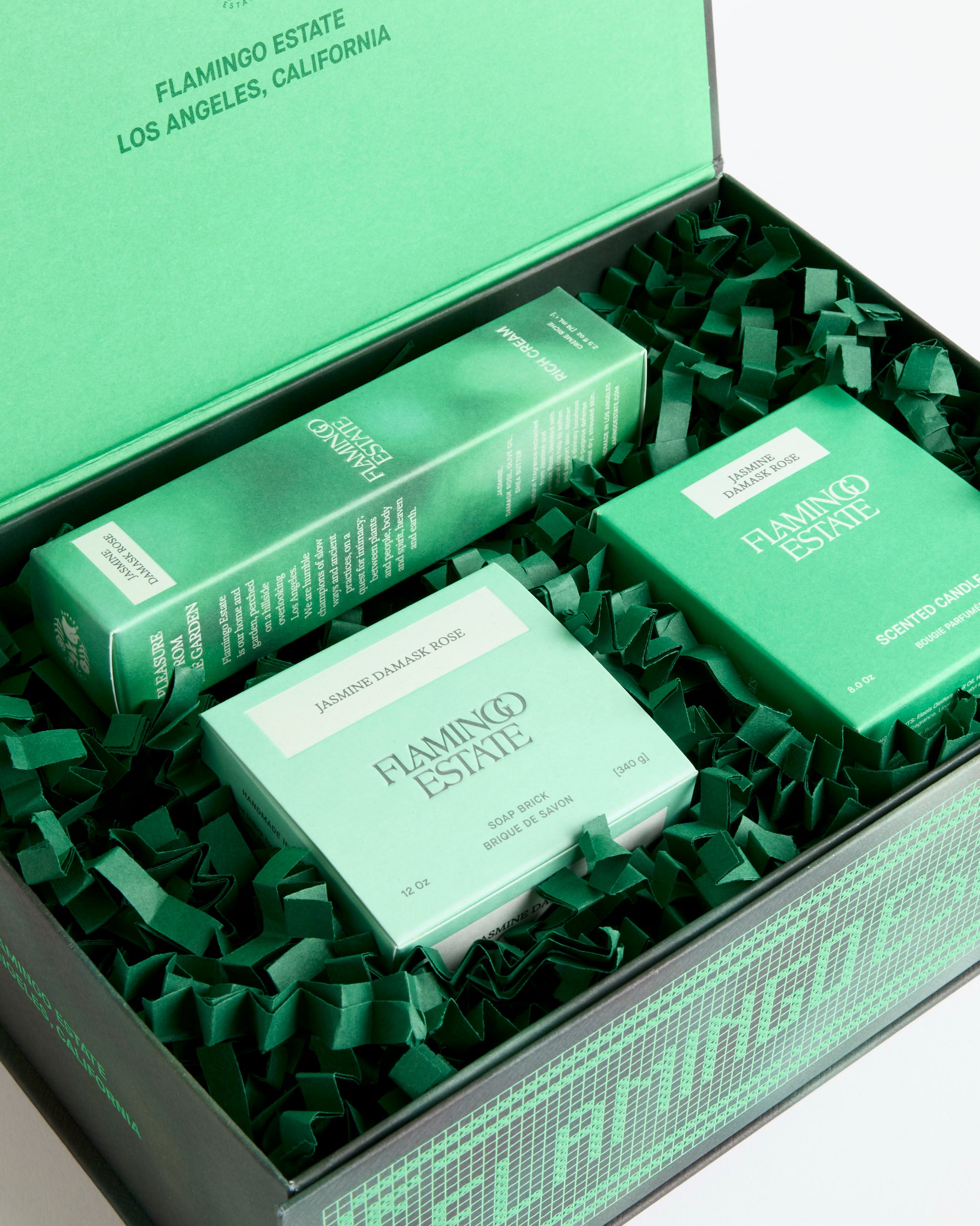 The Jasmine Damask Rose Set by Flamingo Estate includes a scented candle, luxurious Soap Brick, and a boxed item, all nestled on green shredded paper in an elegant green and black Los Angeles gift box.