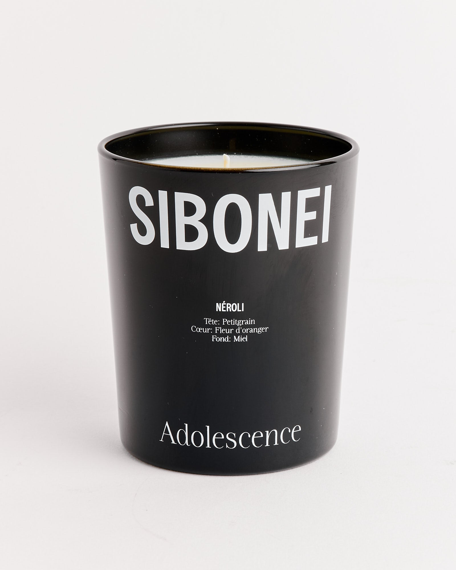A black SIBONEI candle jar labeled “Adolescence” (Neroli) is shown with scent notes of néroli, bergamote, fleur d’oranger, and miel. Made from soy wax, it sits elegantly against a white backdrop.