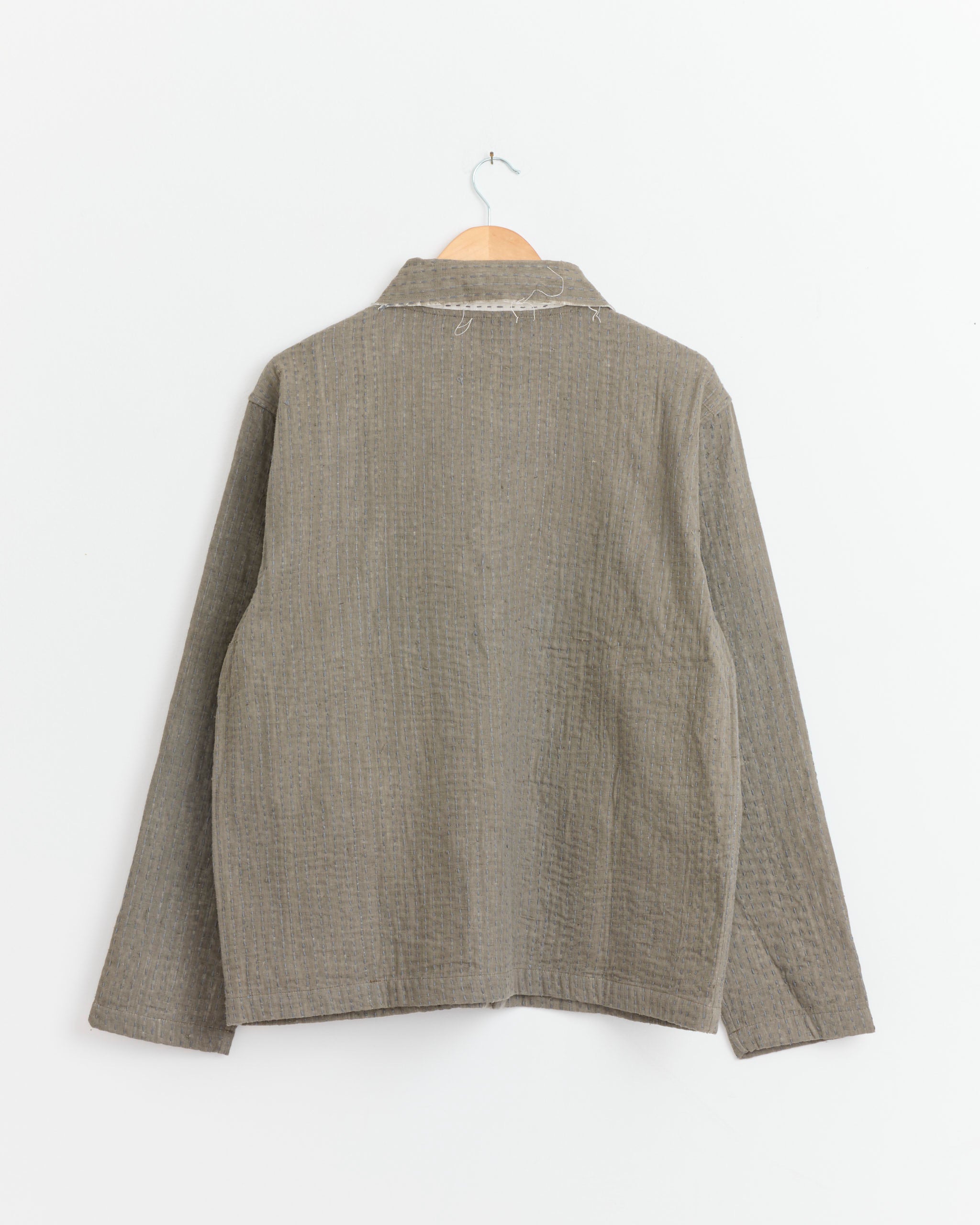 The Kartik Research Kantha Quilt Jacket in Olive, naturally dyed and hand embroidered with long sleeves, hangs on a wooden hanger against a plain white wall, displayed from the back.