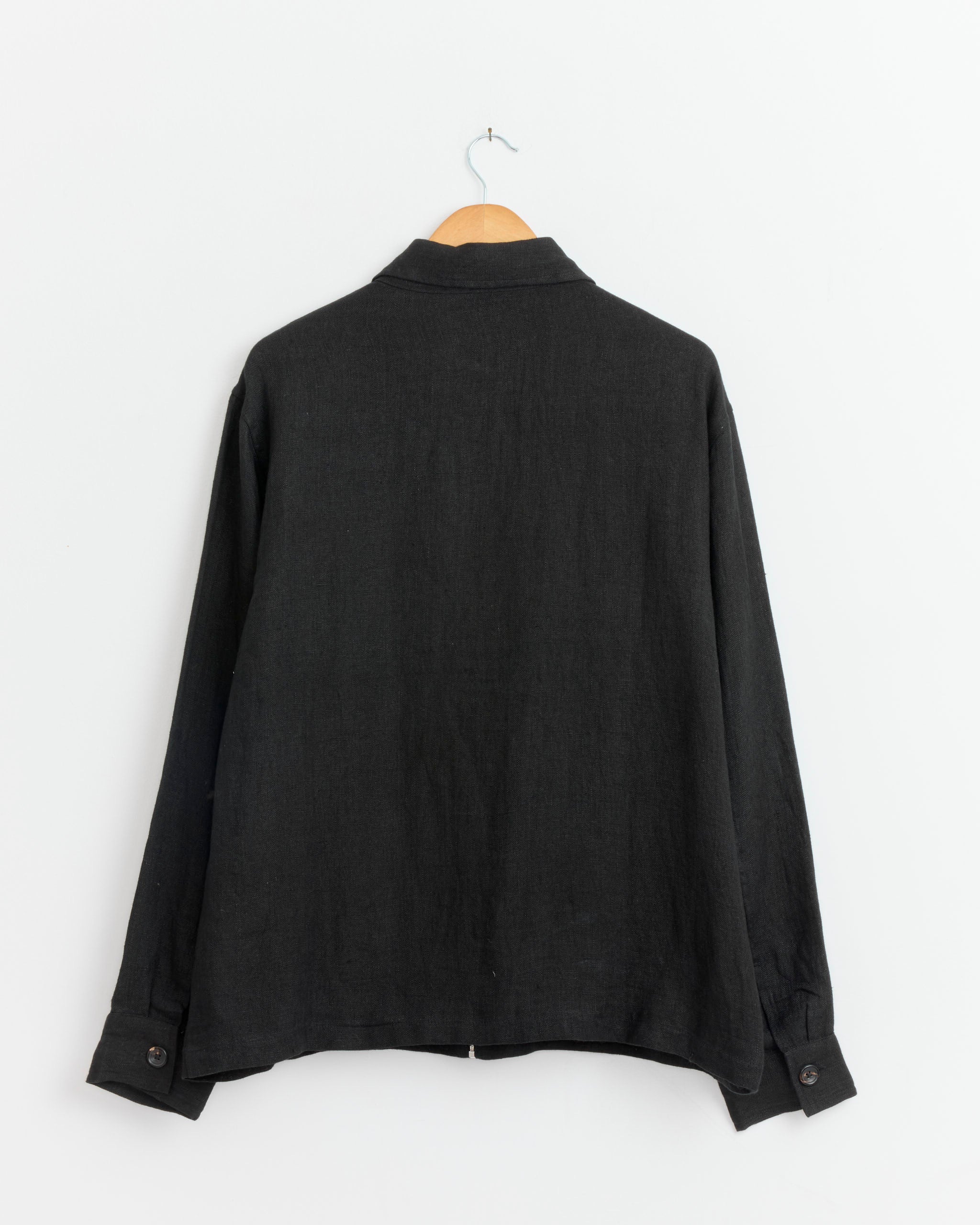 The Kartik Research Zip Work Jacket in Black hangs on a wooden hanger against a white background, shown from the back.