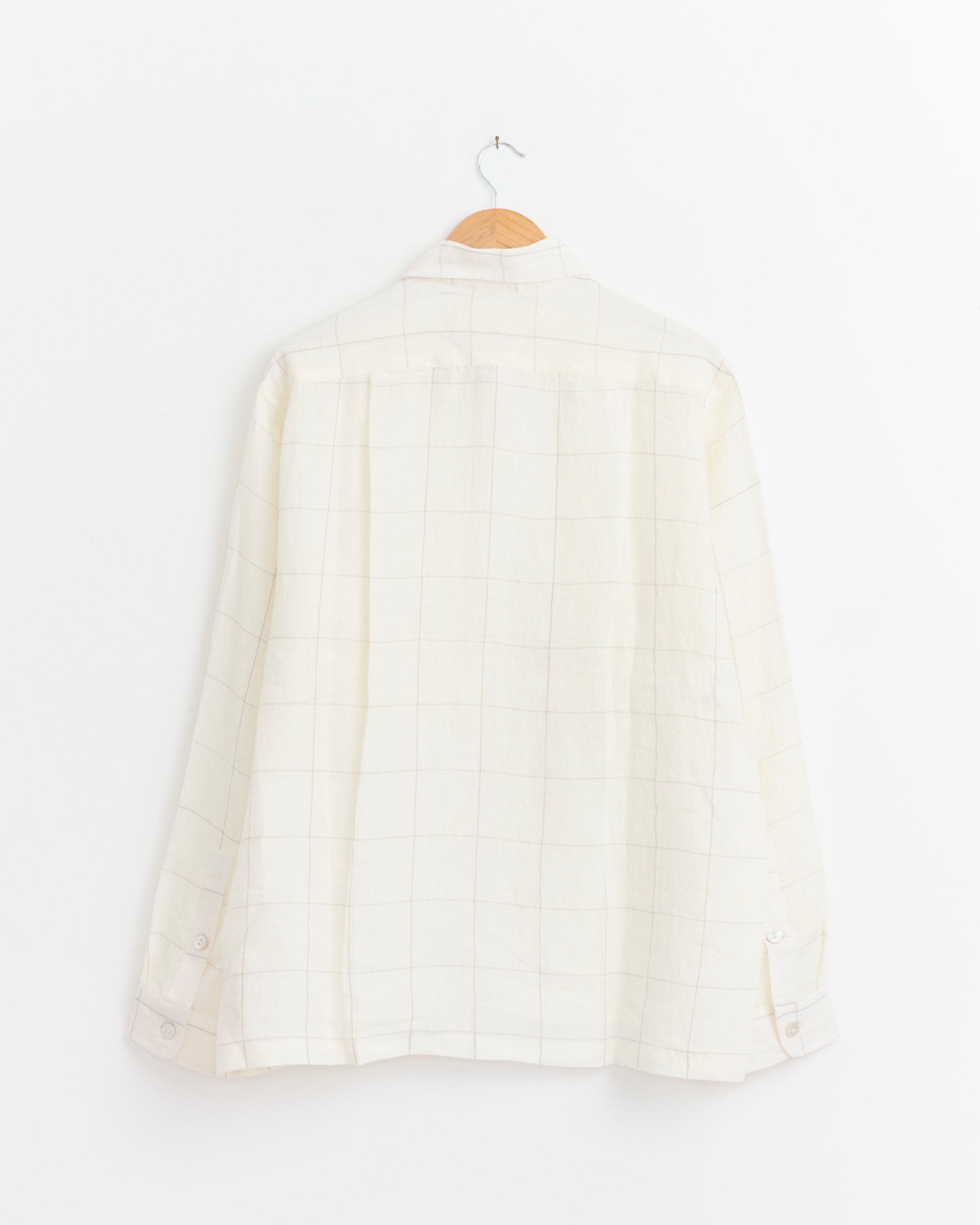 The Pataudi Shirt in Ecru/Silver Zari by Kartik Research, featuring long sleeves and a subtle grid pattern, hangs on a wooden hanger against a plain white wall, displayed from the back.