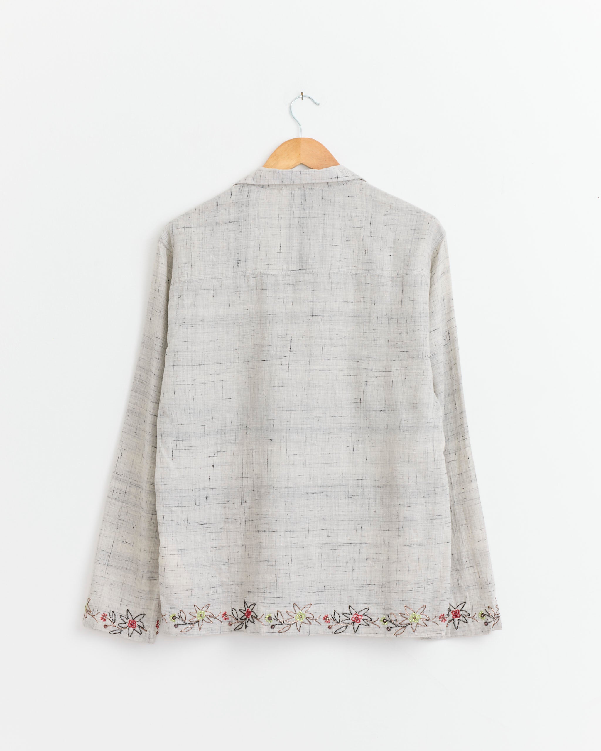 A Beaded Camp Shirt in Grey/Navy/Gold by Kartik Research hangs on a wooden hanger against a white wall, featuring subtle hand-embroidered cotton floral details along the bottom hem.