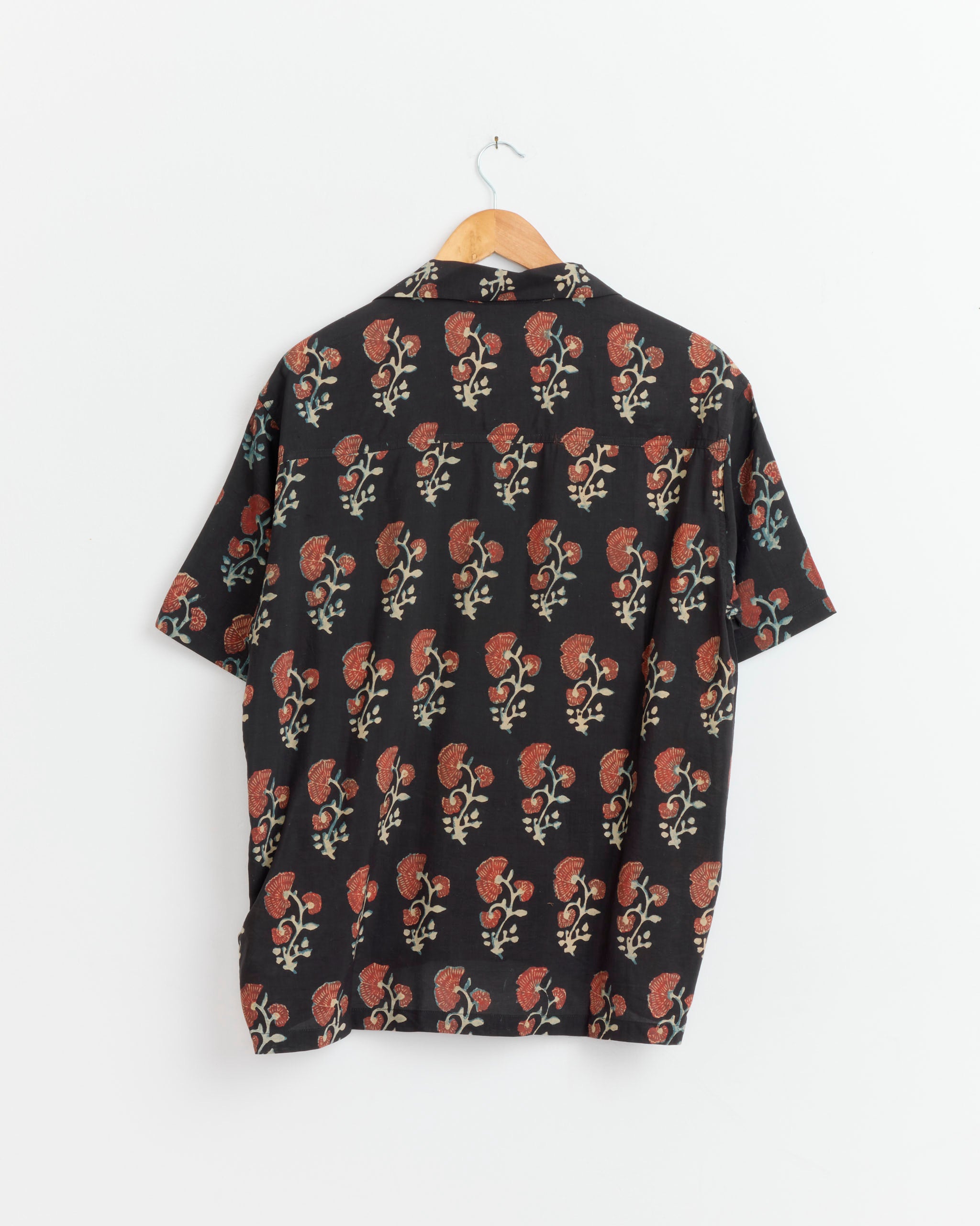 The Kartik Research Camp Shirt in Black/Red/Yellow features a short-sleeve, button-up design with a floral paisley motif, displayed on a wooden hanger against a white wall.