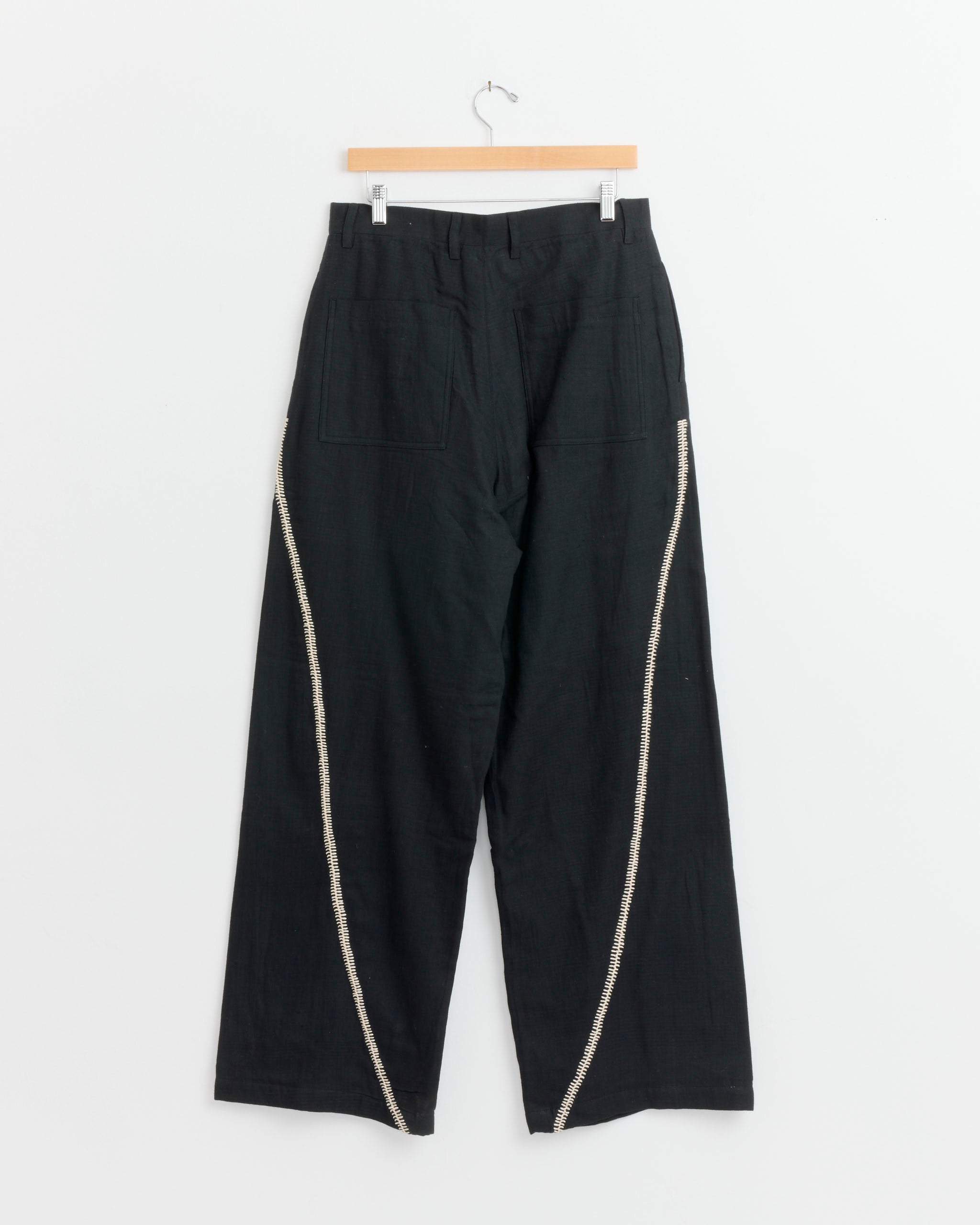 The Rabari Sultan Pant in Black/Ecru by Kartik Research is a pair of black wide-leg handwoven wool pants with two back pockets and light-colored decorative side stitching, shown hanging on a wooden hanger.