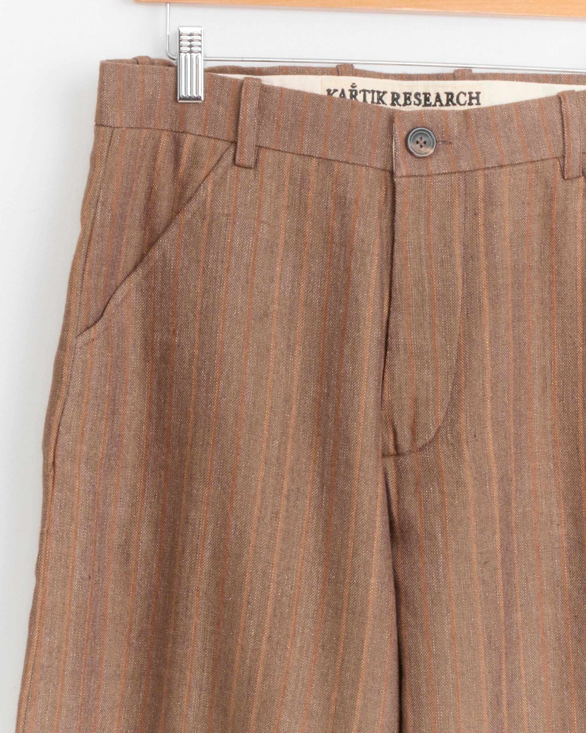 Close-up of the Flat Front Flare Trouser in Purple/Brown by Kartik Research, made in India, hanging on a wooden hanger. The image highlights the waistband, button, zipper, front pockets, and inner brand label.