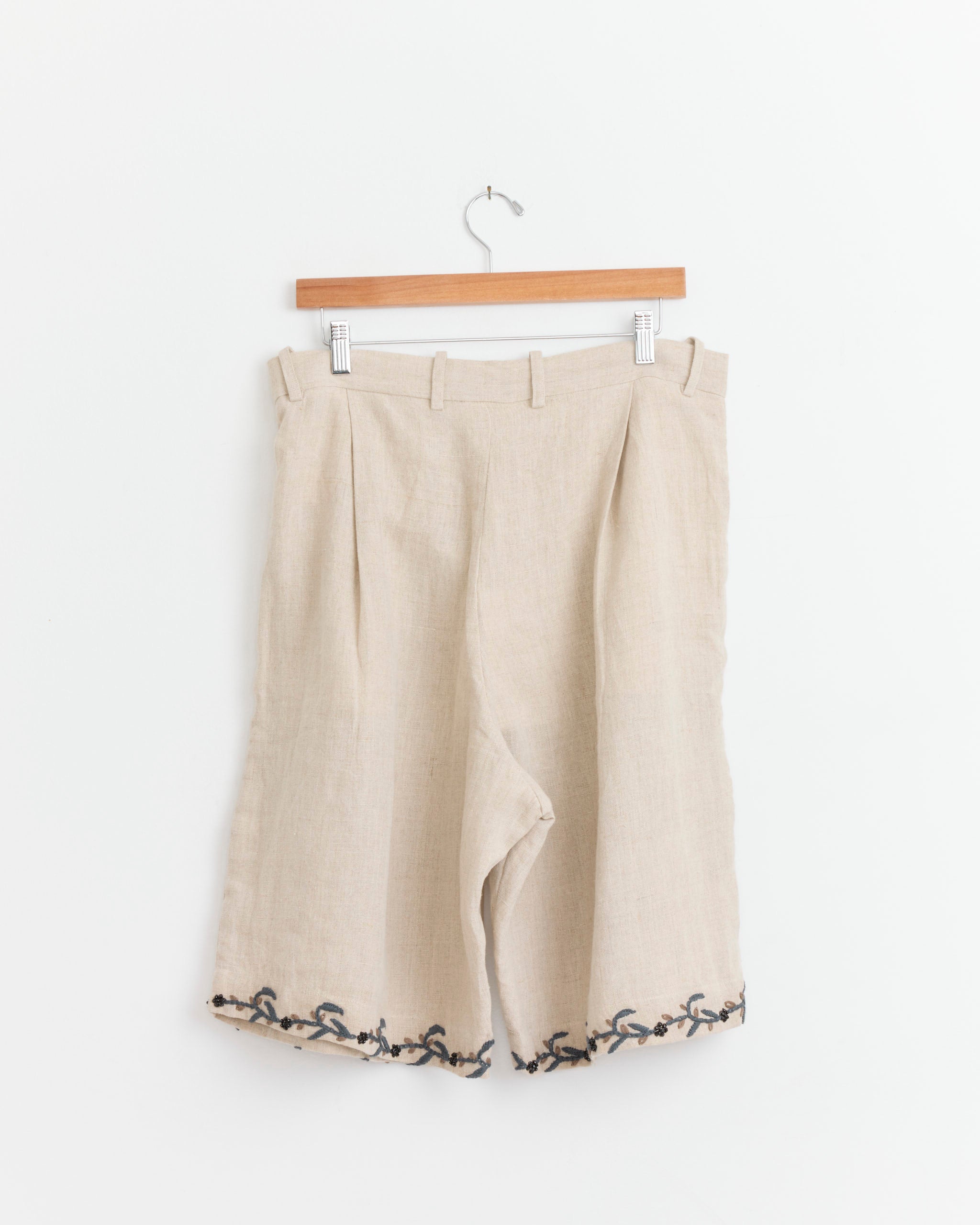 The Kartik Research Big Short in Beige/Black/Grey features handloom linen, belt loops, pleats, and black embroidery along the hem. Made in India, these shorts are shown hanging on a wooden hanger.