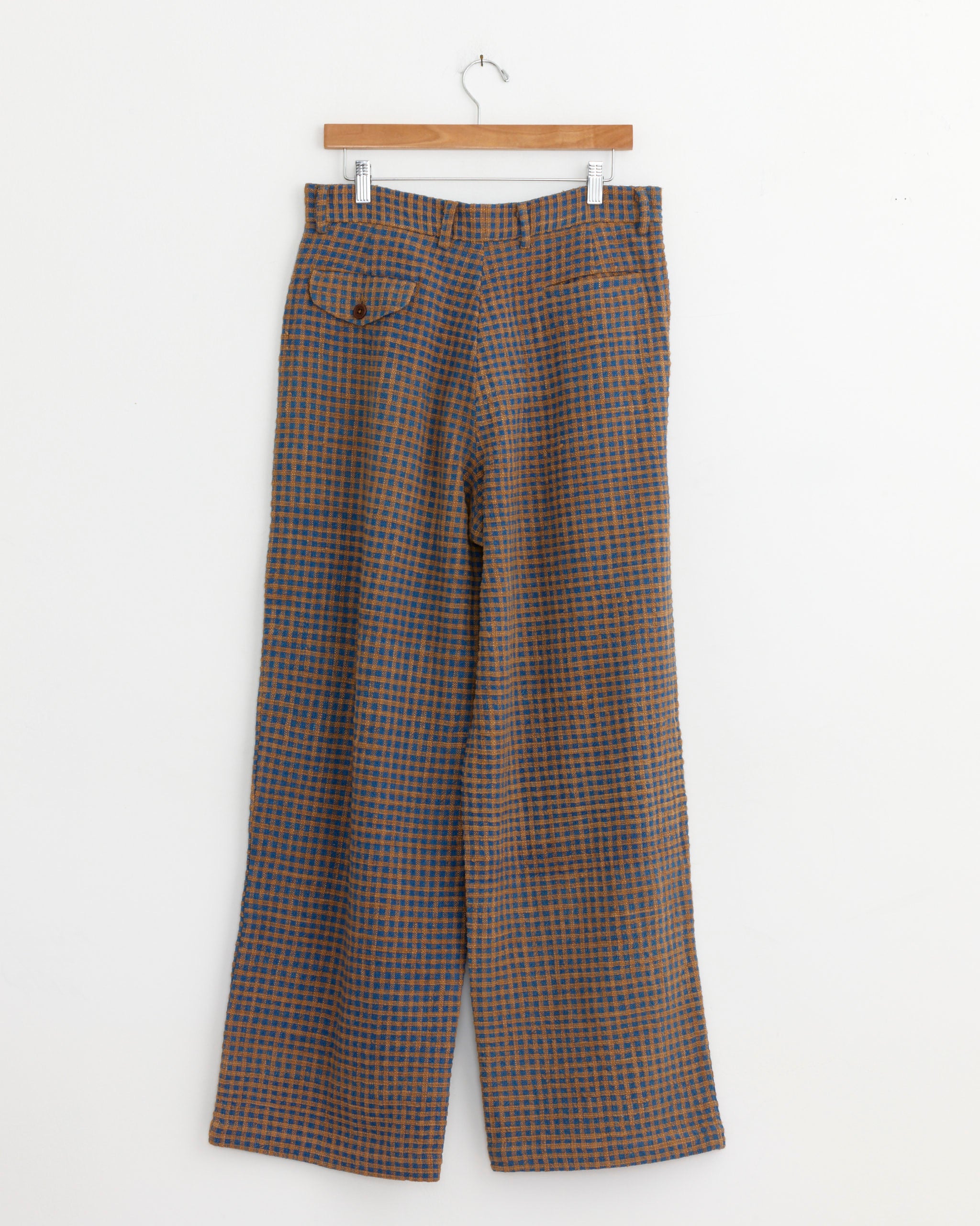 The Story mfg Fenn Trouser in Wobbly Waffle is a pair of high-waisted, wide-leg organic cotton trousers with a blue, brown, and orange checkered pattern, shown on a wooden hanger against a plain white wall.