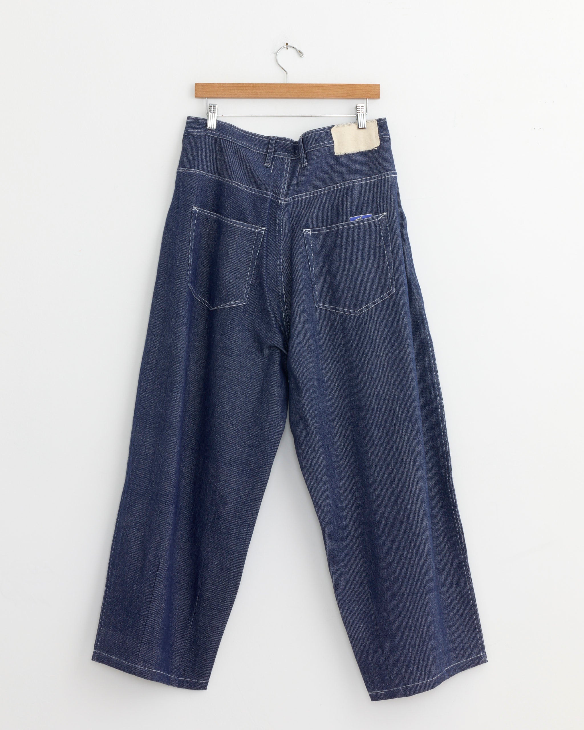 The Story mfg Lush Jean in Earthloom Handloom Denim, vintage-inspired wide-leg blue jeans with two back pockets and a light fabric patch near the waistband, hangs on a wooden hanger against a plain white wall.