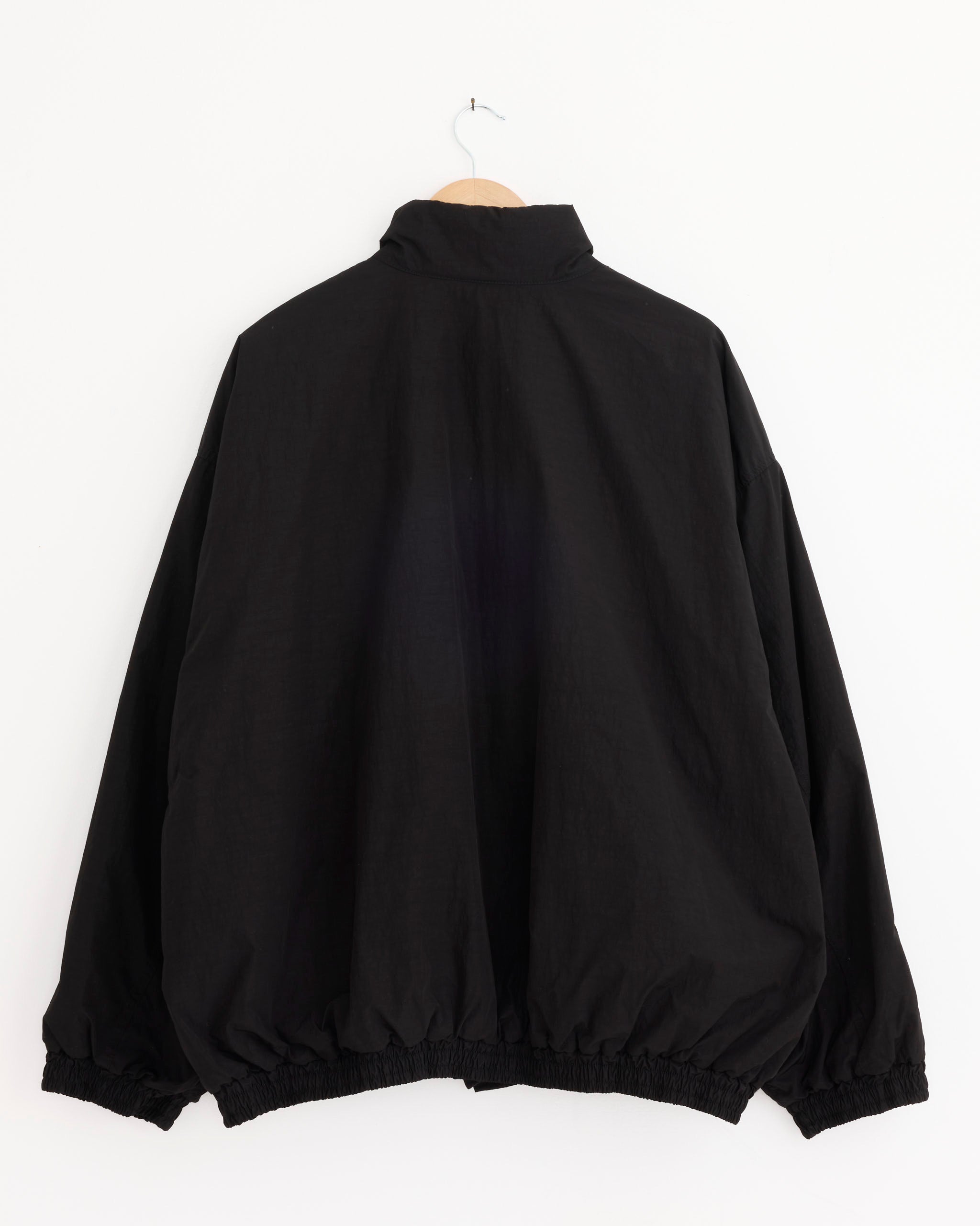 The ssstein 1375 Jacket in Black, featuring a high collar plus elastic cuffs and hem, is made in Japan. It hangs on a wooden hanger against a plain white wall, shown from the back.