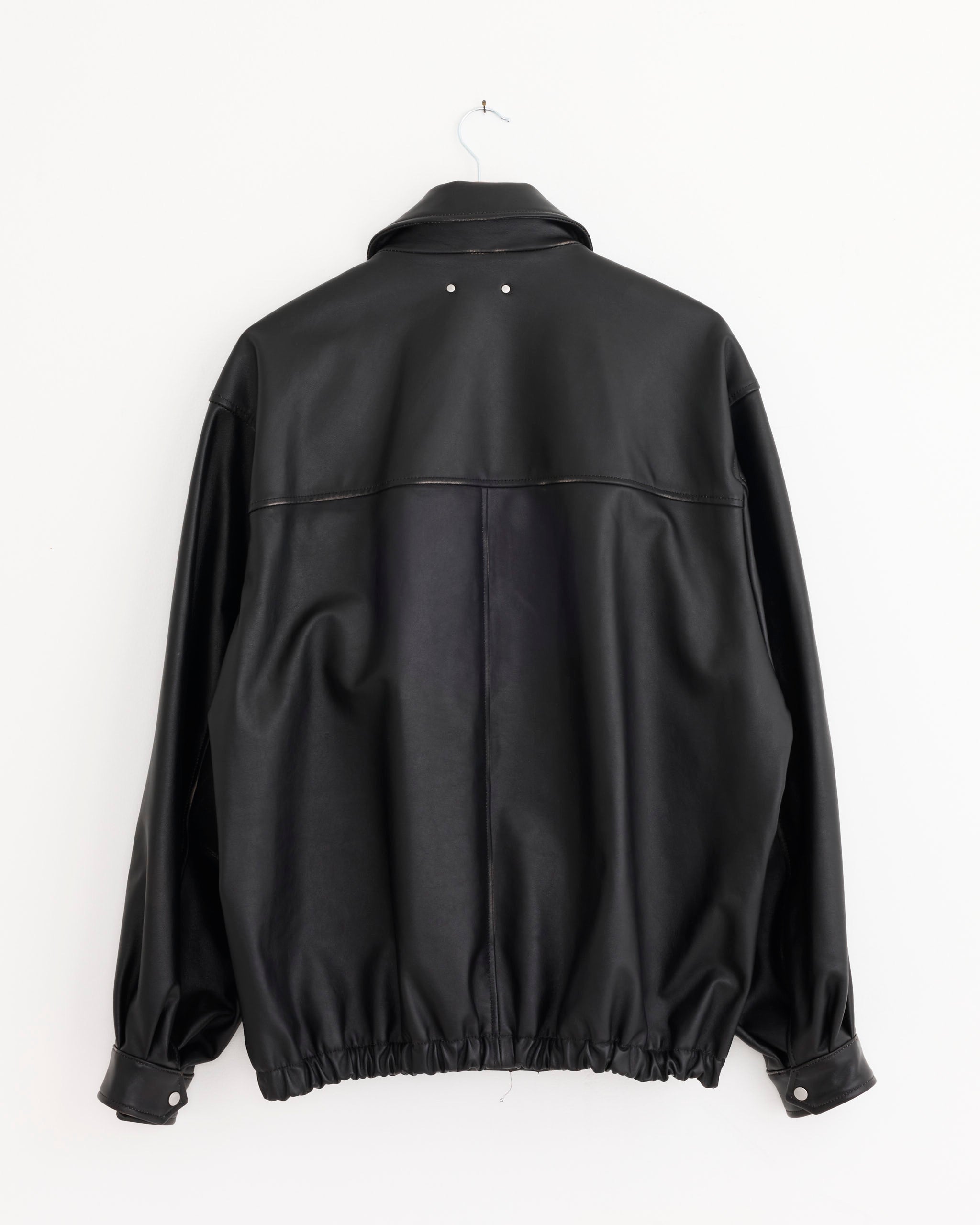 The ssstein 1380 Jacket in Black, made of cow leather, hangs on a white wall, shown from the back. It features a relaxed fit with gathered cuffs and waistband, plus two small silver rivets near the collar.