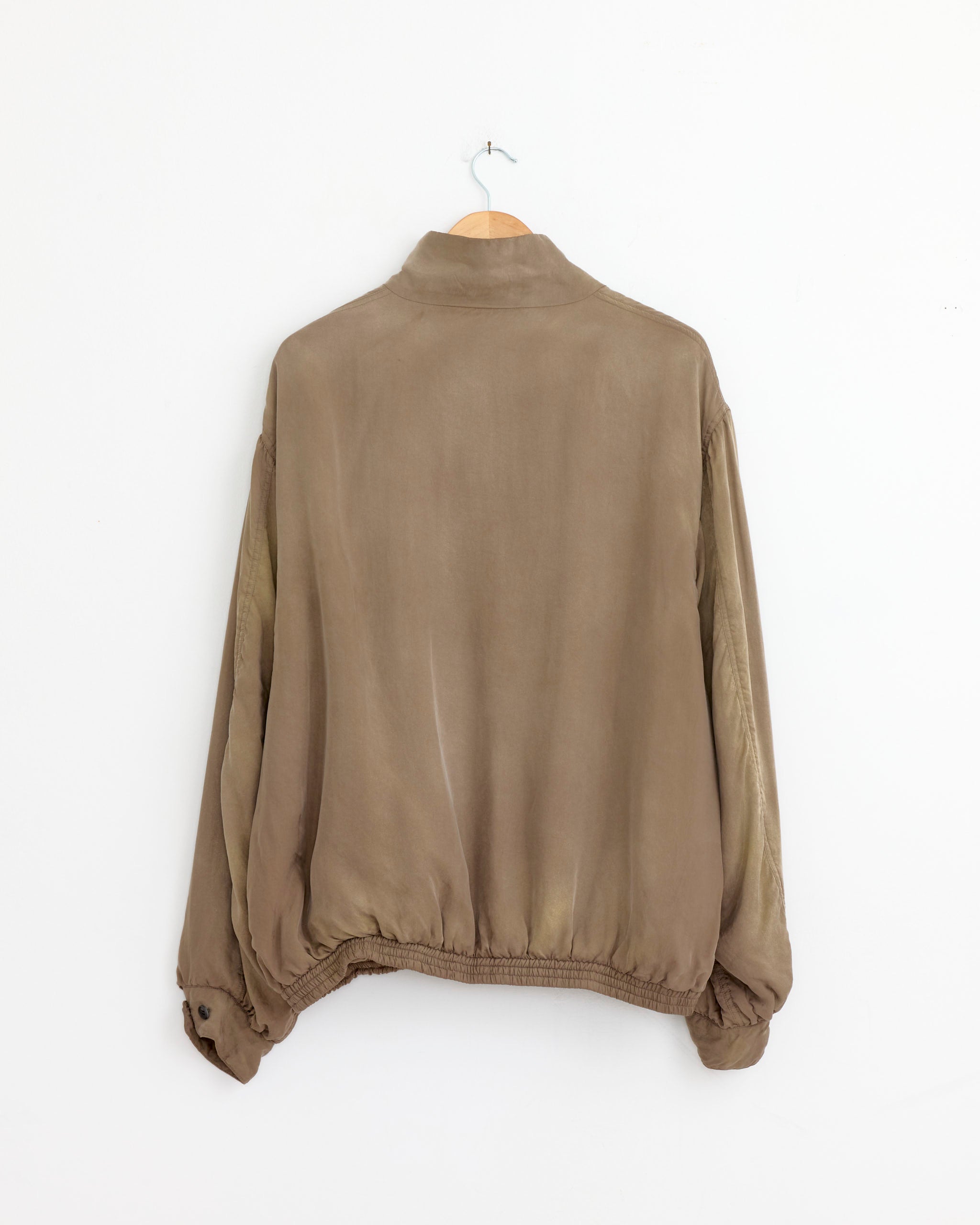 The ssstein 1388 Zip Jacket in Khaki, featuring long sleeves and a vintage look in light brown, hangs from a wooden hanger against a plain white wall, displayed from the back.