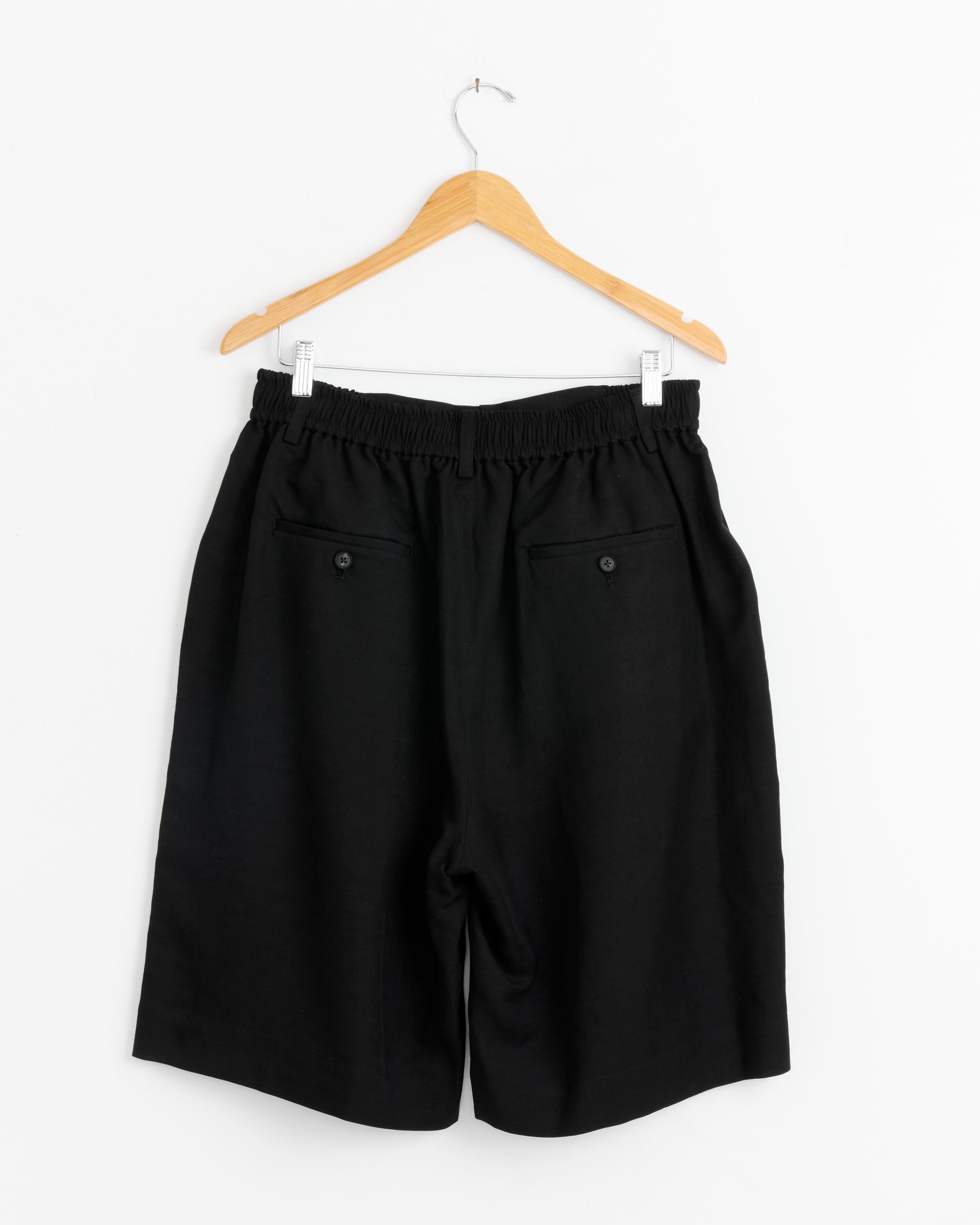 The ssstein 1437 Short in Black features a wide, long-length cut, elastic waistband, and two buttoned back pockets. Made in Japan from lightweight rayon-linen twill, it's shown on a wooden hanger against a plain white background.