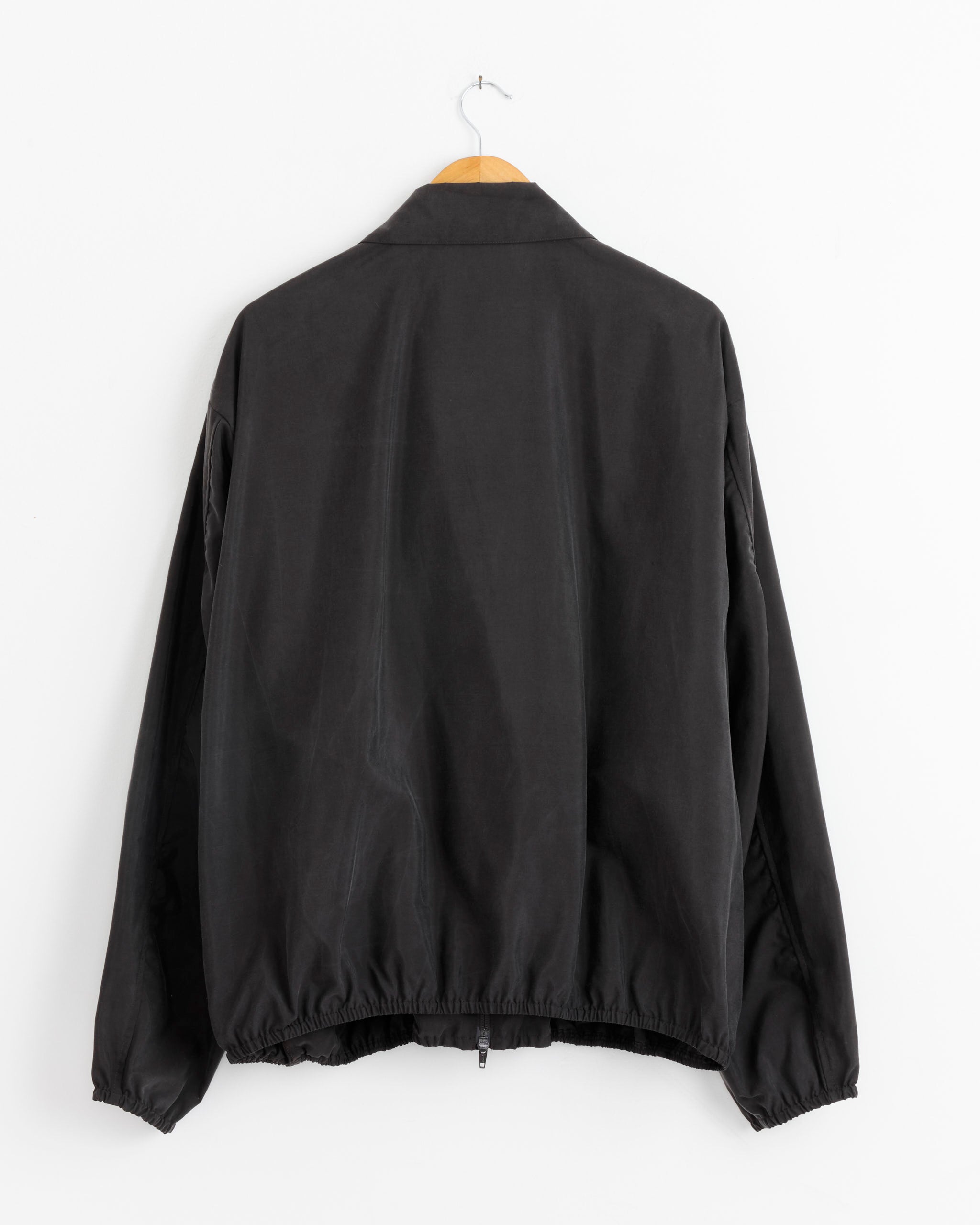 A brown ssstein 1442 Jacket with long sleeves, made in Japan, hangs on a wooden hanger against a white wall, shown from the back.