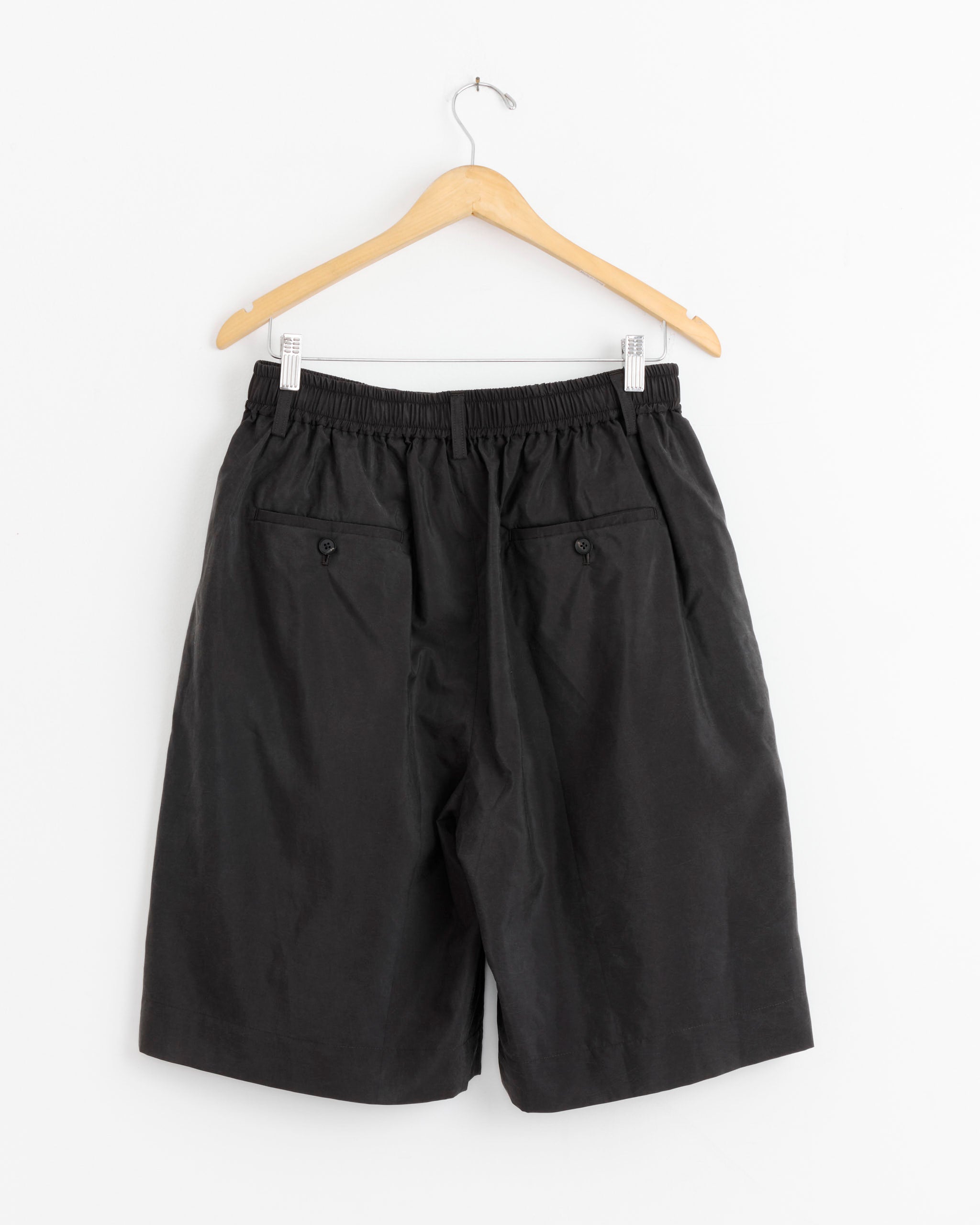 The ssstein 1446 Short in Brown, featuring a relaxed fit, elastic waistband, and buttoned back pockets, hangs on a wooden hanger against a white wall.