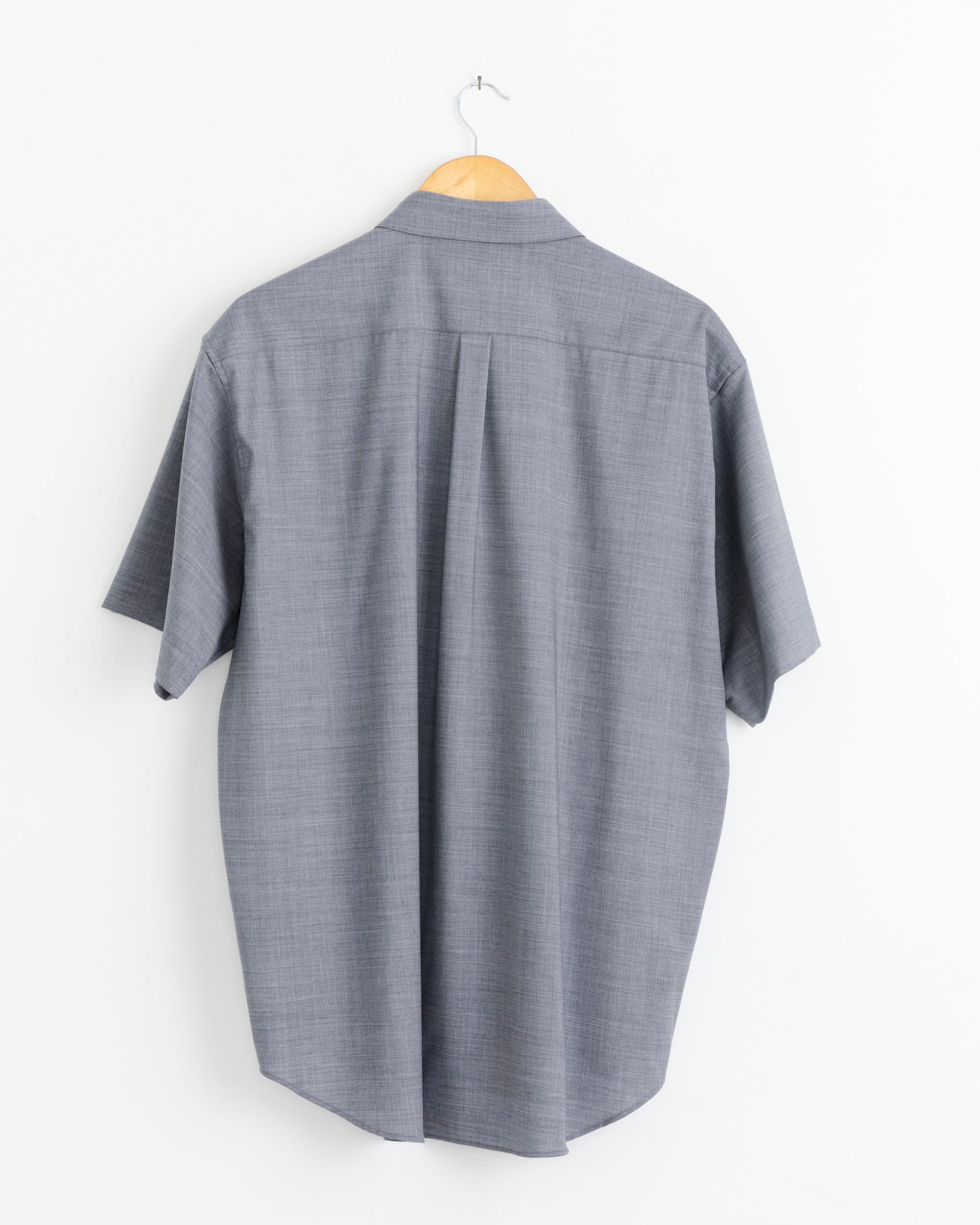A blue ssstein 1499 Shirt, crafted in Japan from luxurious Super100’s wool, hangs on a wooden hanger against a plain white wall, shown from the back.