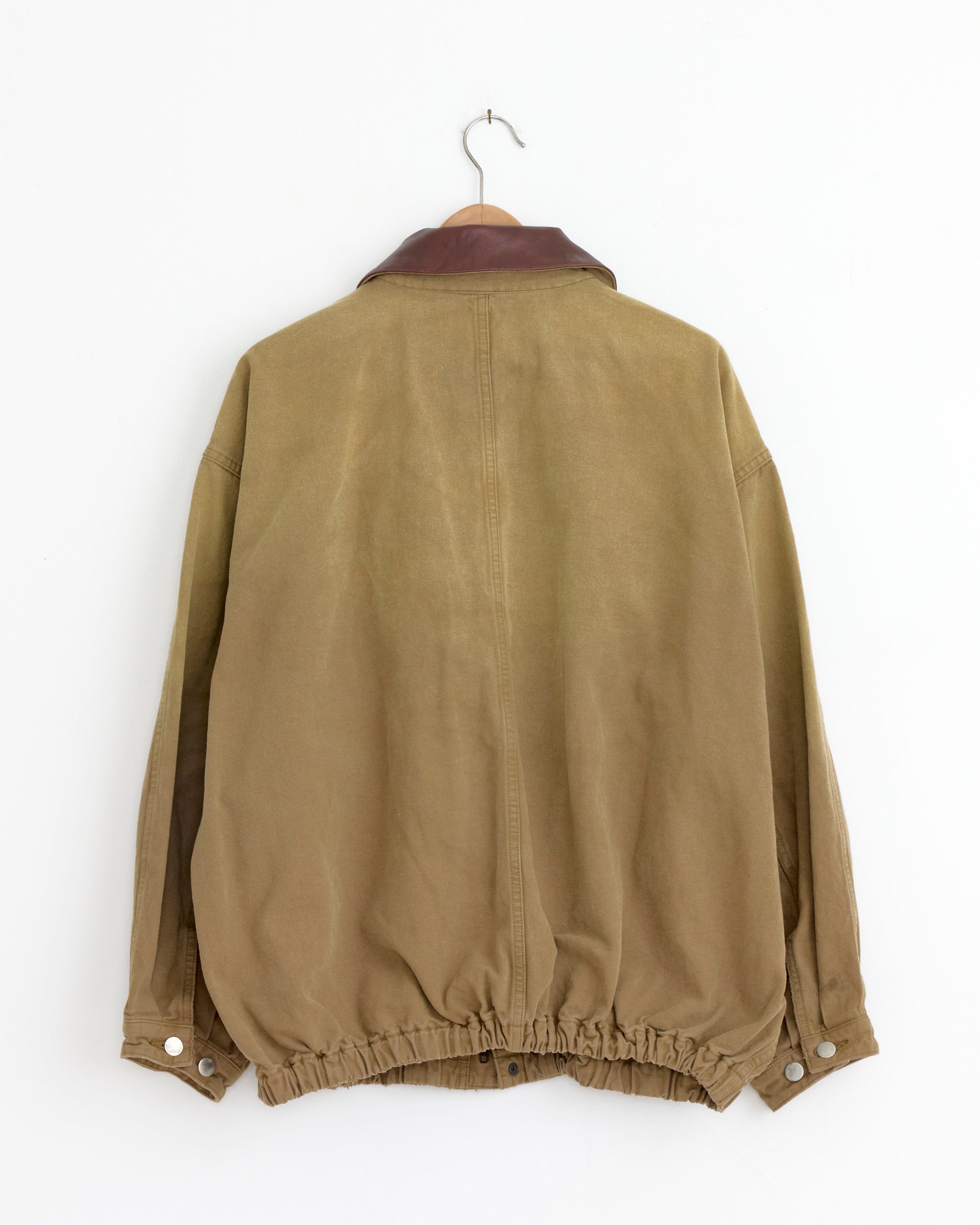 The ssstein 1387 Jacket in Khaki Beige hangs on a hook, shown from the back against a plain white wall. It features an oversized fit, brown collar, elasticated waist and cuffs, and button closures.