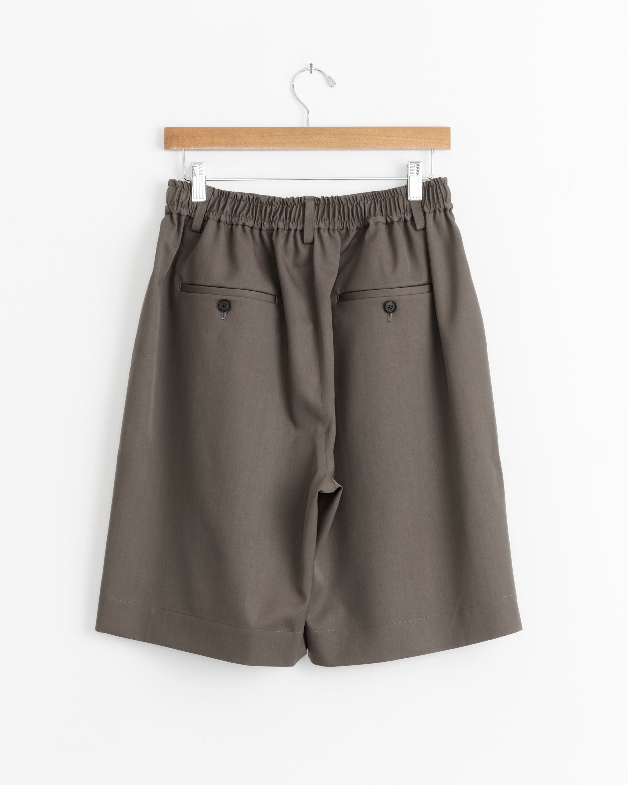 The ssstein Wide Easy Short in Dark Beige features a loose fit, knee-length pleats, an elastic waistband, and two buttoned back pockets, displayed on a wooden hanger against a white background.
