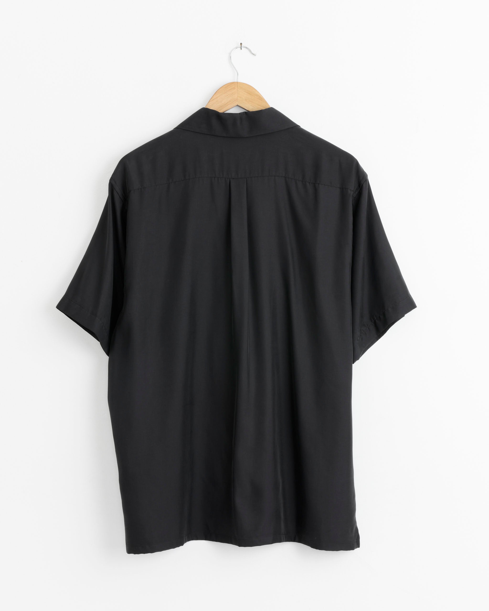 The ssstein Cupro Twill Oversize Shirt in Black, made in Japan, hangs on a wooden hanger against a white wall, displayed from the back with short sleeves.