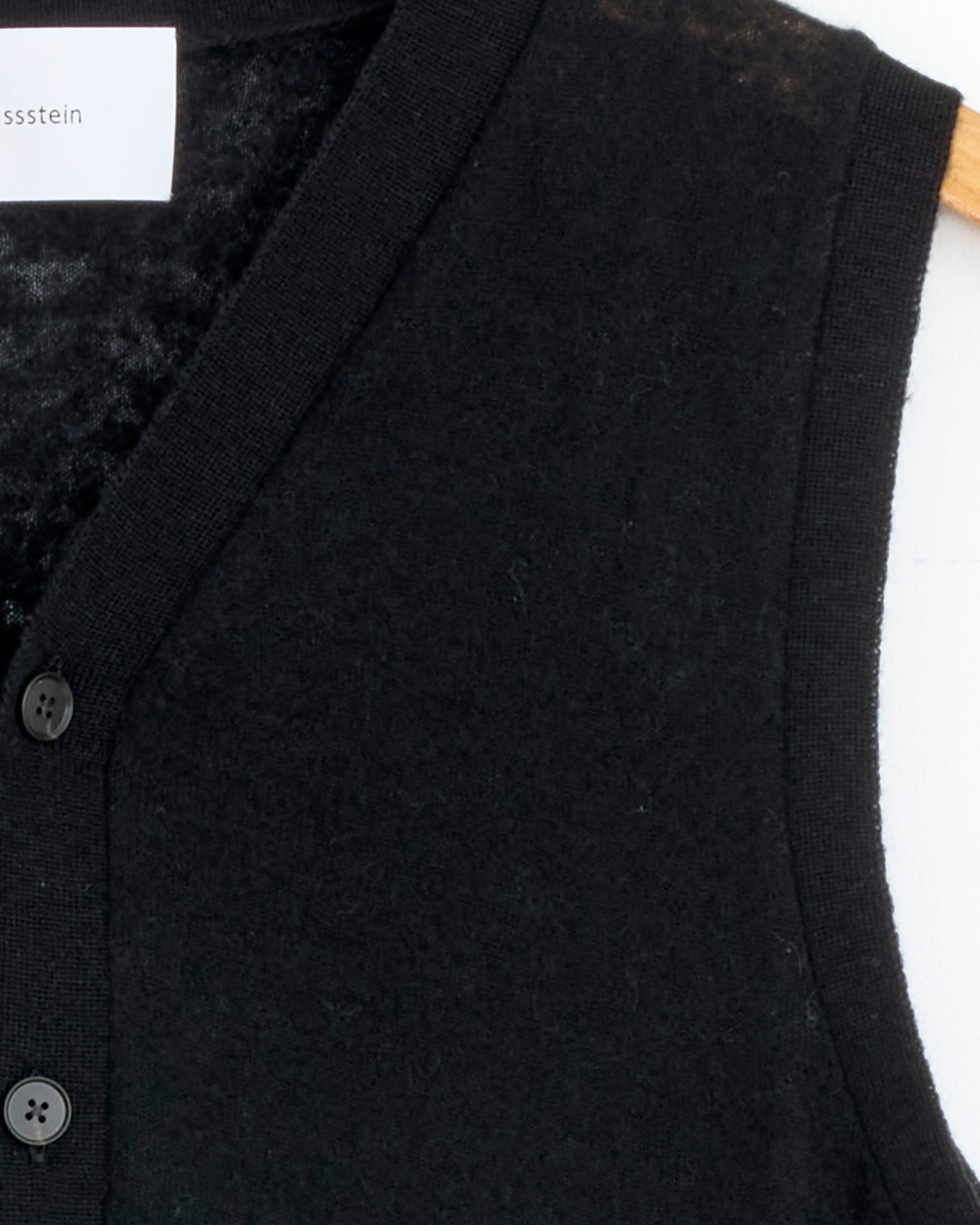 Close-up of the upper left side of the ssstein 1482 Vest in Black, a sleeveless knit vest made in Japan, shown on a hanger with part of a white label visible inside the collar.