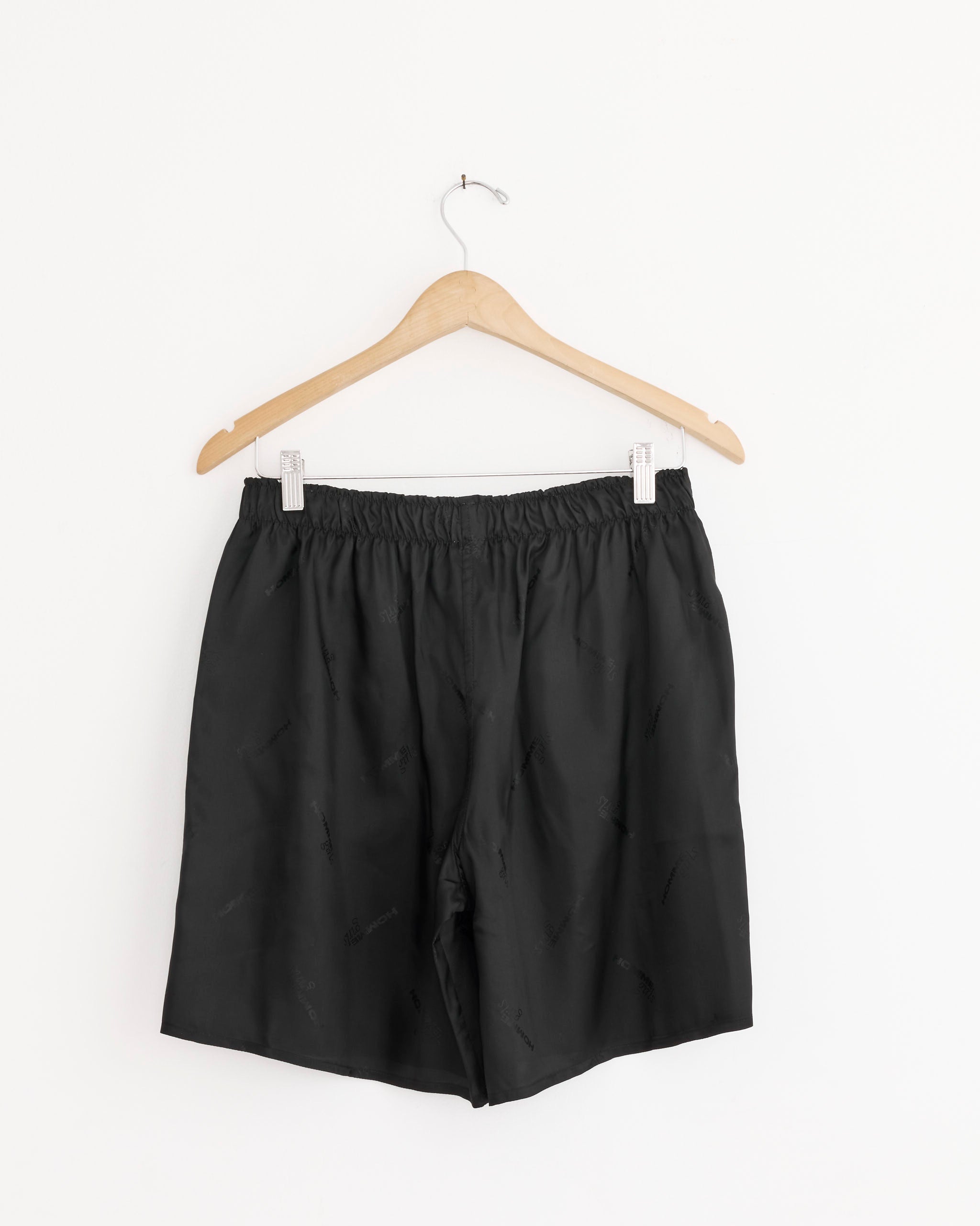 The Hommegirls Long Boxer Short in Black Logo—black satin shorts with an elastic waistband—hangs on a wooden hanger against a plain white background.