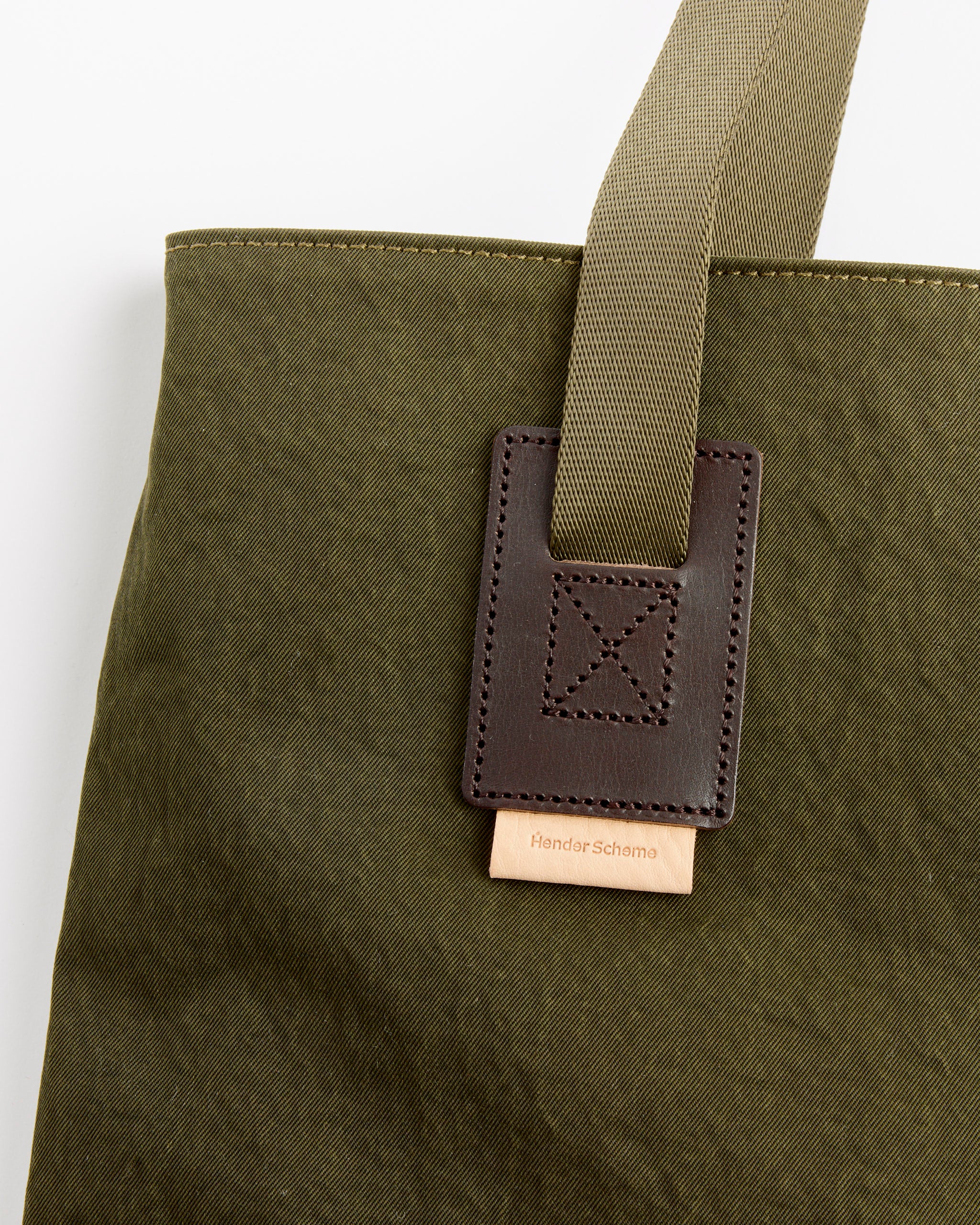 Close-up of the Two Way Tote in Khaki Green by Hender Scheme, featuring a tan woven handle and brown leather accent. A small leather tag with the brand name is stitched beneath the handle accent. For sizing questions, contact us via online chat.