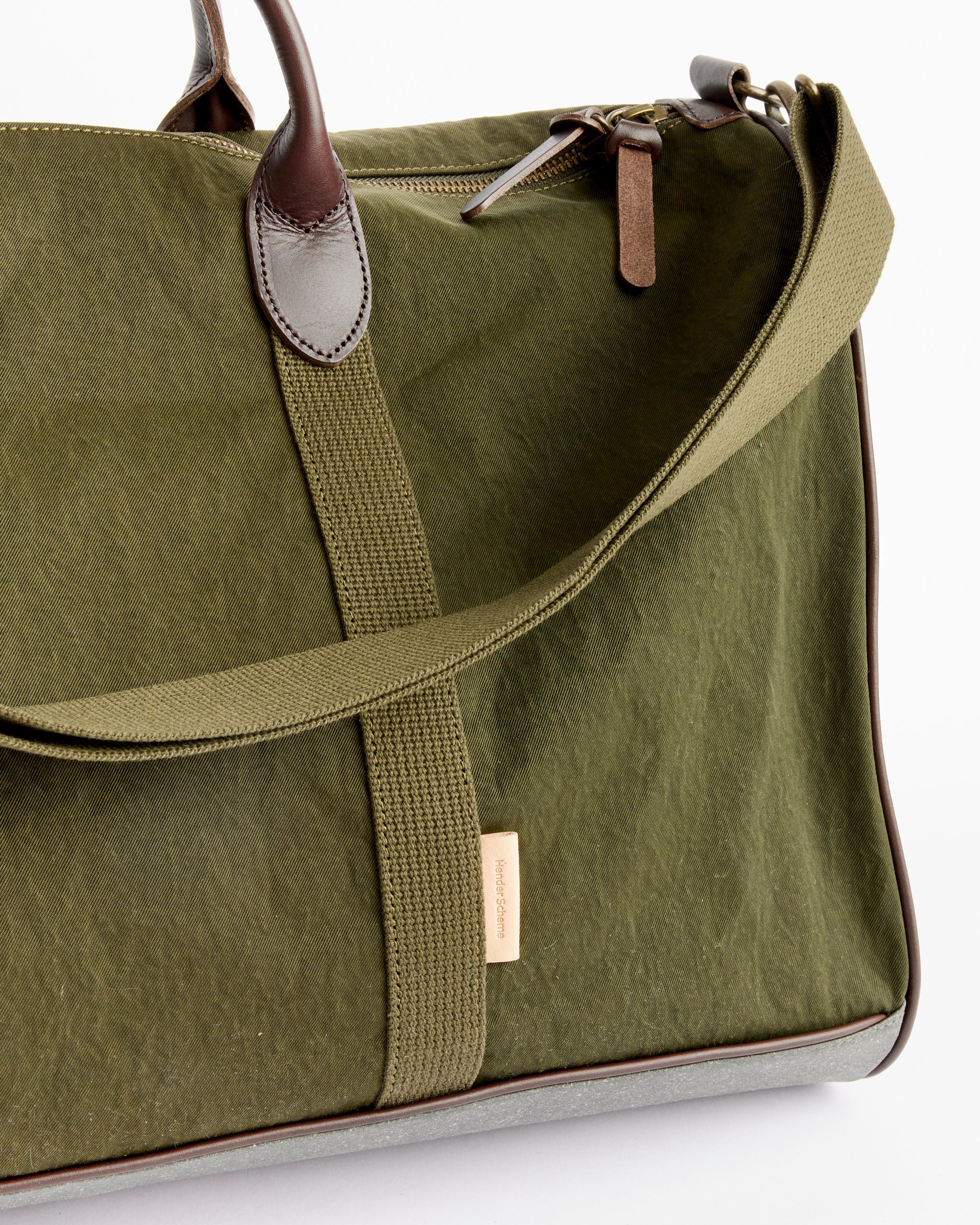 A close-up of the Hender Scheme Boston Luggage in Khaki Green shows brown leather handles, a matching shoulder strap, a beige tag, and visible zipper and stitching details.