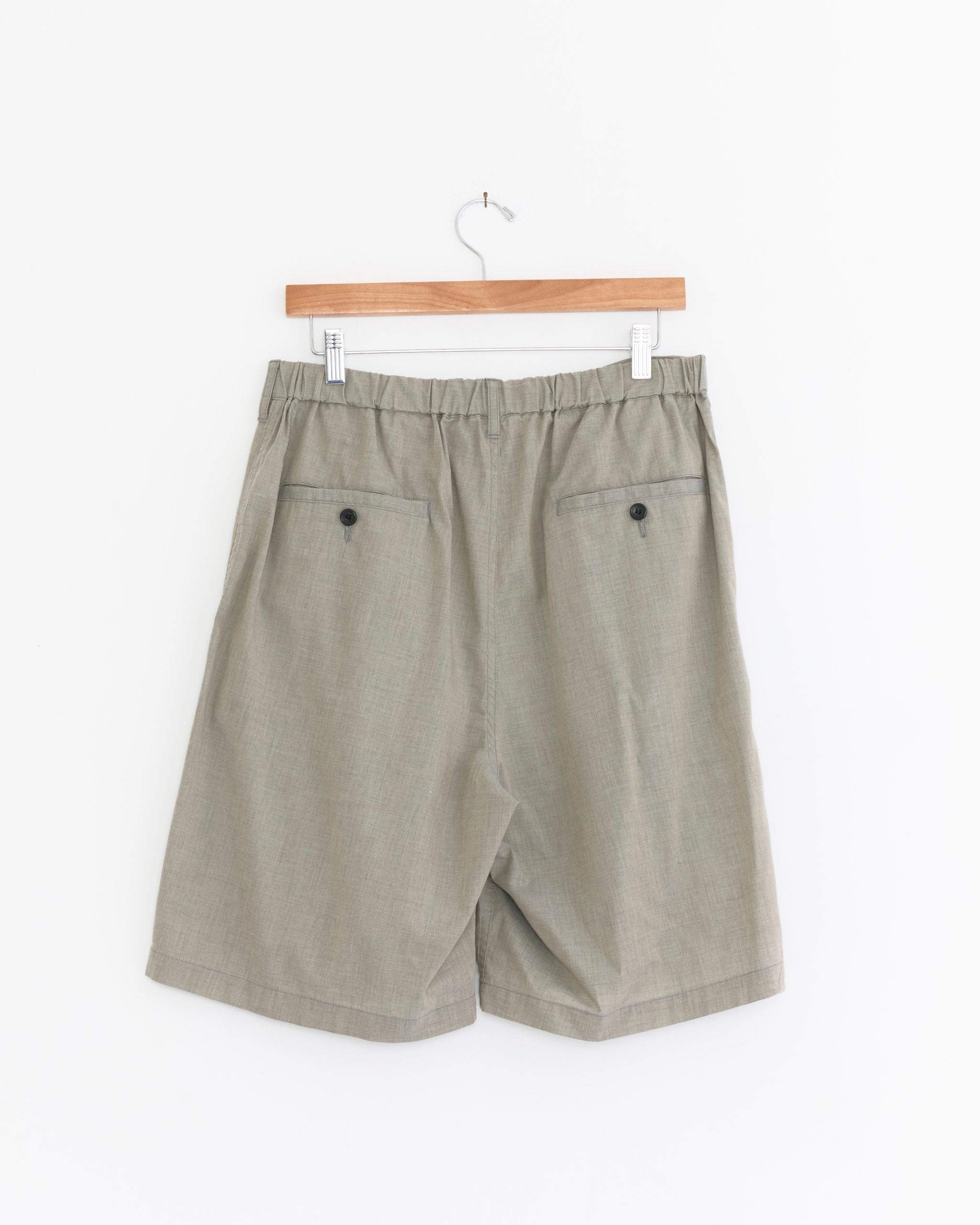 The Graphpaper Cotton Wool Poplin Oversized Short in Grey, made in Japan, features an elastic waistband and two buttoned back pockets, shown hanging on a wooden hanger against a plain white background.