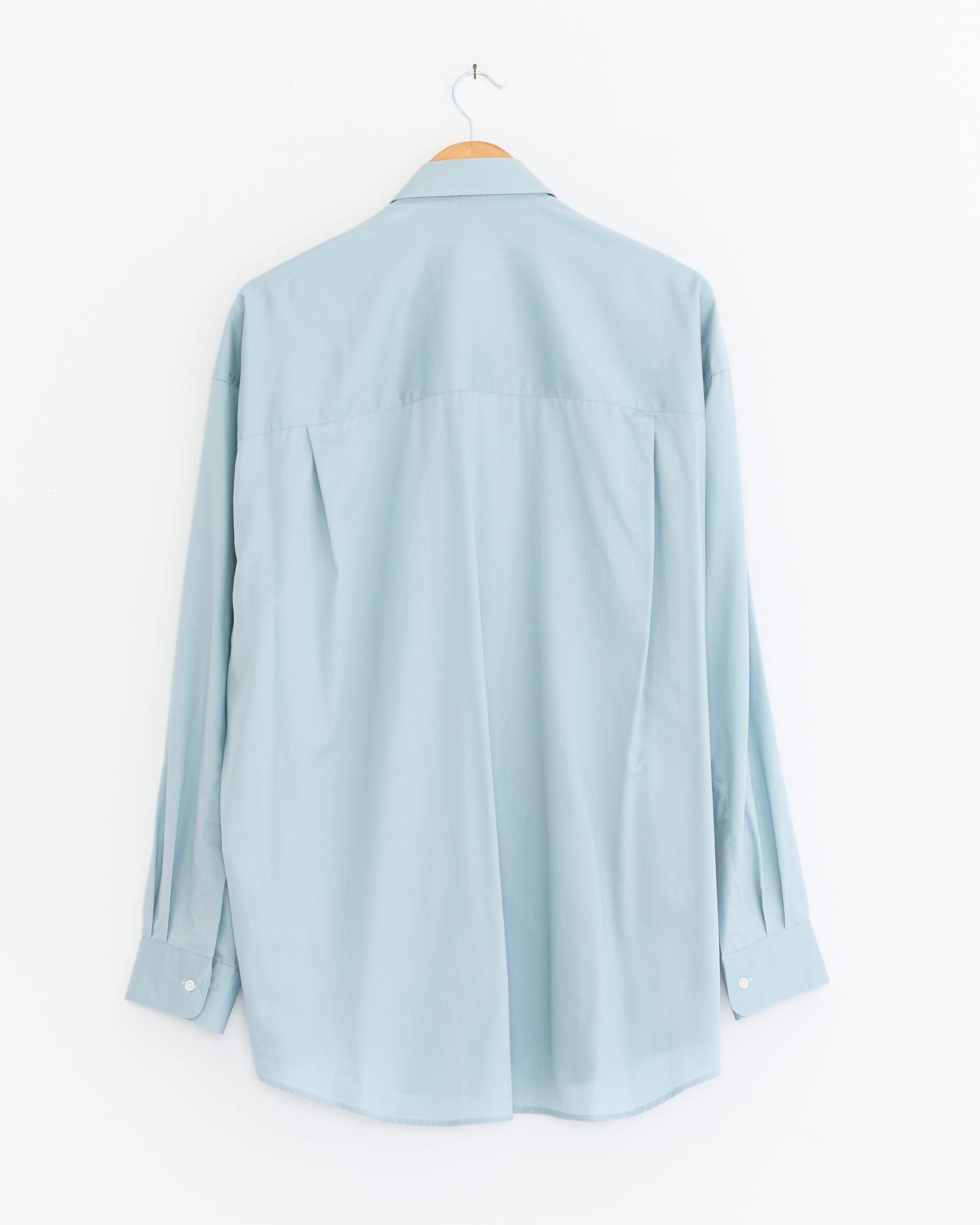 The Graphpaper Silicon Poplin Oversized Regular Collar Shirt in Smoked Blue is displayed from the back, long-sleeved and hanging on a wooden hanger against a plain white background.