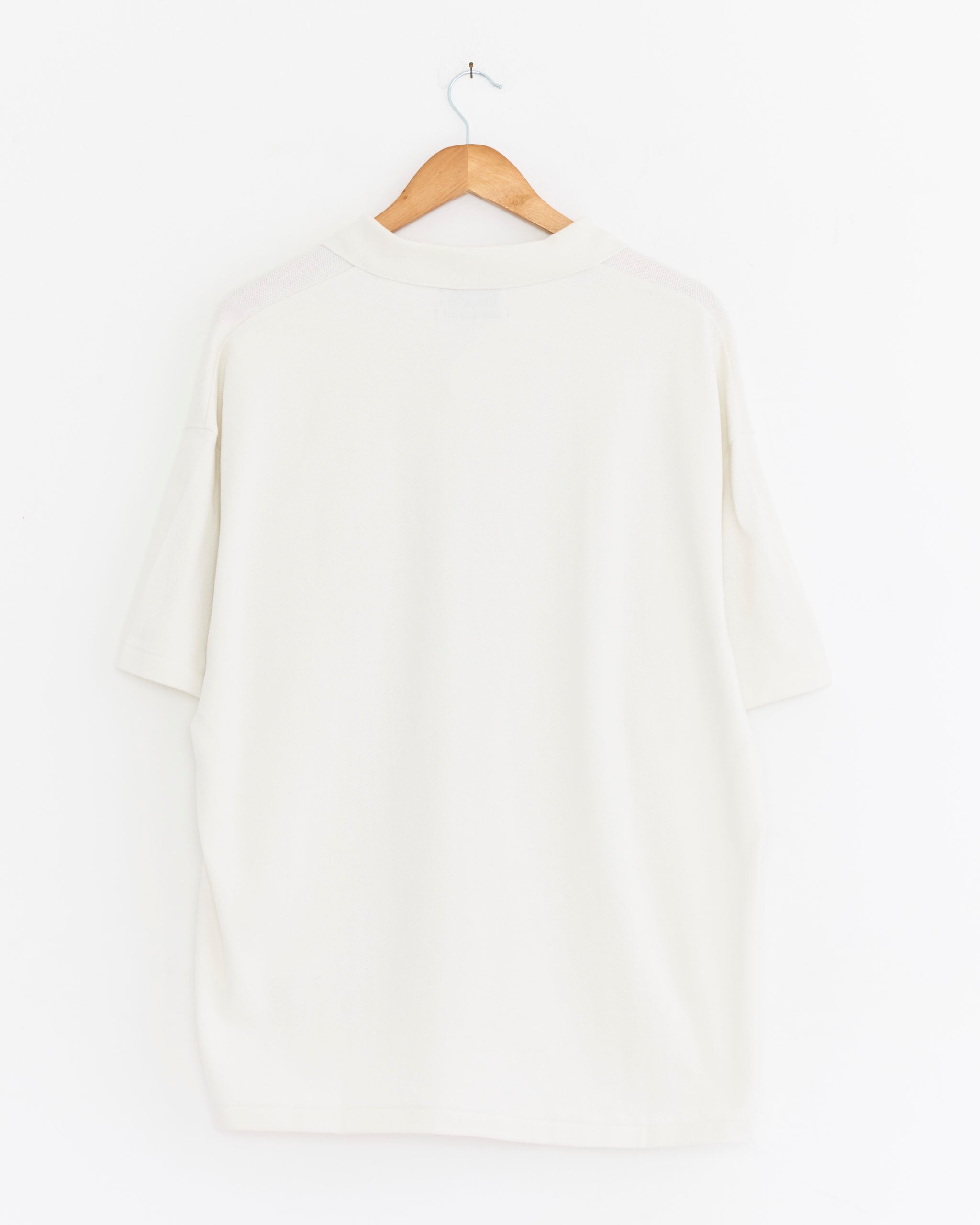 The Graphpaper Cotton Pique Jersey Skipper Polo in Pearl White, featuring a relaxed fit and short sleeves, is displayed from the back on a wooden hanger against a white background.