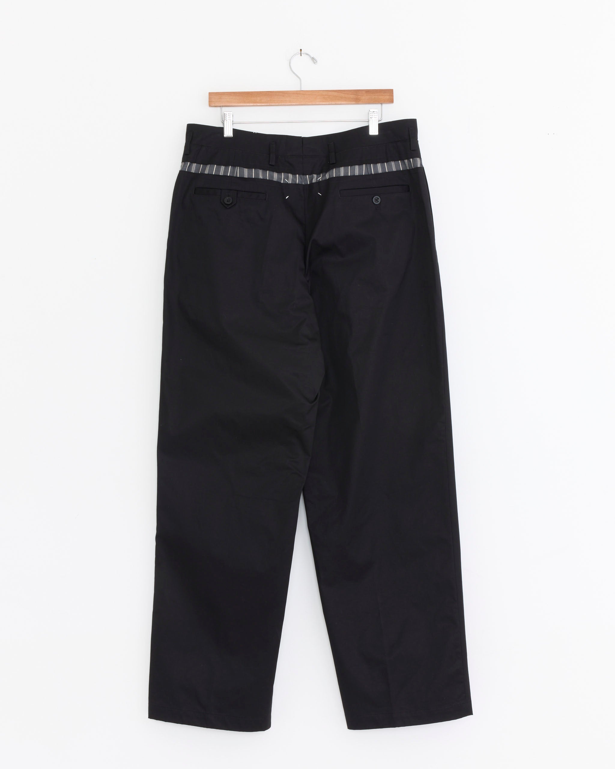 Maison Margiela’s Waxed cotton Wide-Leg Trousers in Black hang on a wooden hanger against a white backdrop, displayed from the back with a checkered waistband and buttoned back pockets.