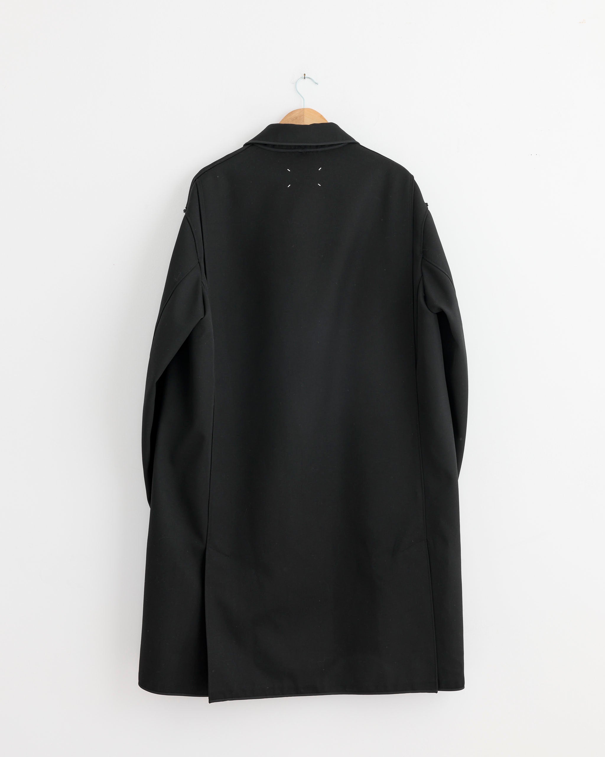 A Maison Margiela Reversible Coat in Black/Grey Check hangs on a wooden hanger against a white wall, displayed from the back to highlight its minimal, modern design.