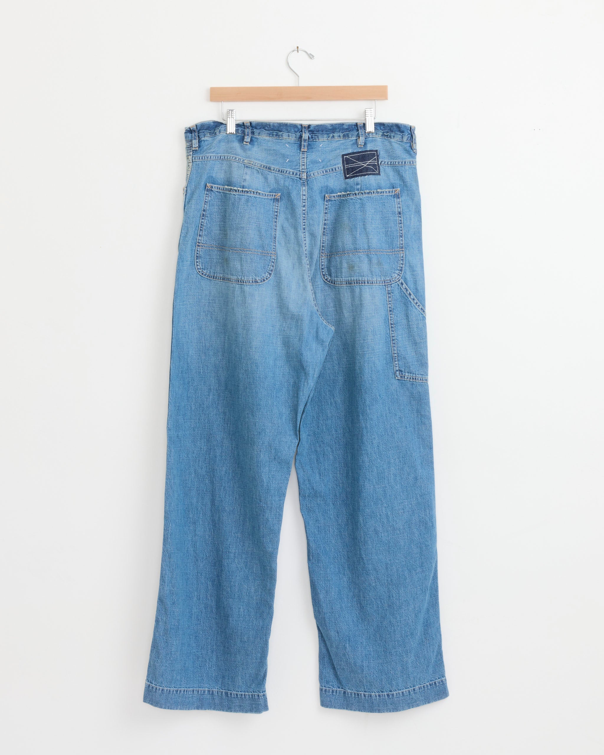 Maison Margiela's Pant in Blue Washed features wide-leg denim in a light wash, shown from the back with two large pockets and a leather waistband patch, displayed on a wooden hanger against a white background.