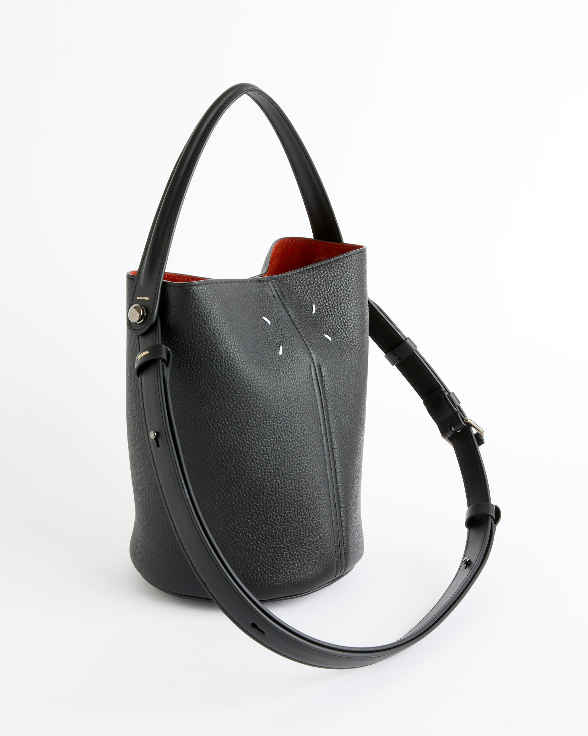 The Dress-Age Hobo Bag by Maison Margiela is crafted in black pebbled calfskin with a single top handle, adjustable shoulder strap, red interior lining, white stitched accents on the front, and subtle equestrian-inspired details.