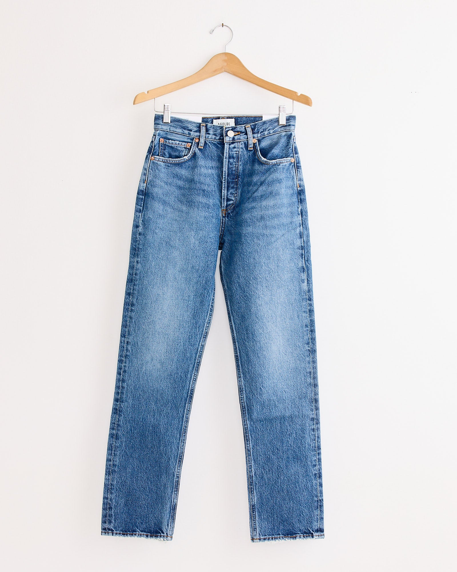The AGOLDE 90's Pinch Waist Jean in Essence, made from regenerative cotton denim with a classic straight-leg fit, hangs on a wooden hanger against a plain white wall.