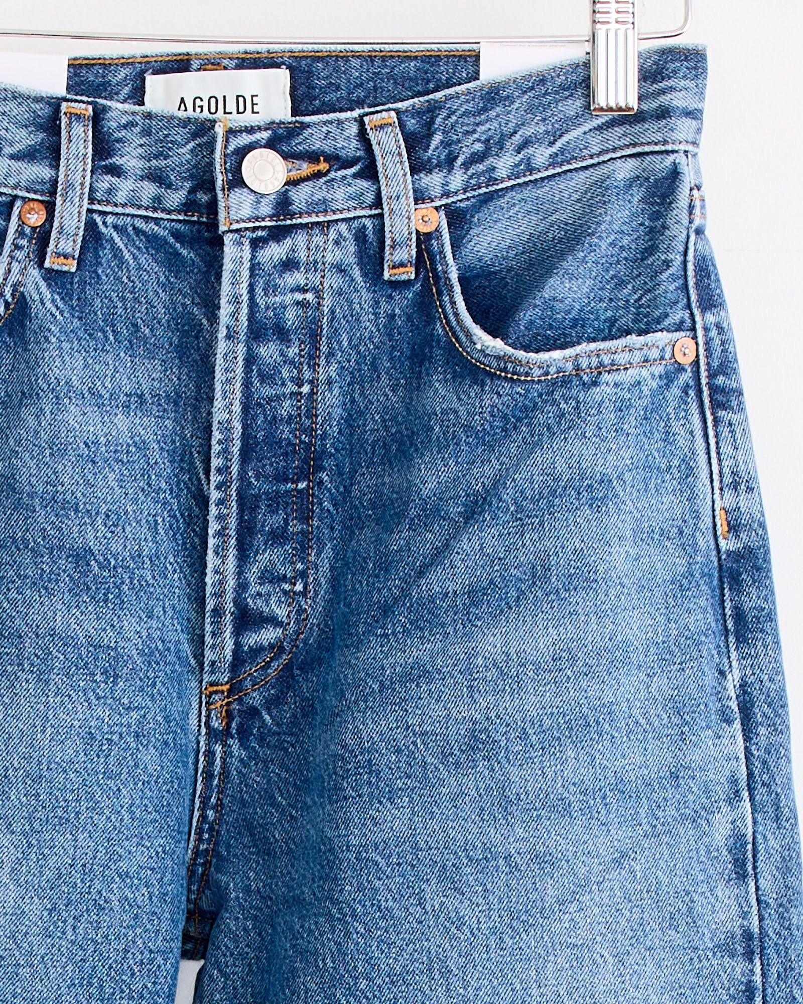 Close-up of AGOLDE’s 90's Pinch Waist Jean in Essence, blue regenerative cotton denim with a high-rise straight leg, copper rivets, front button, belt loops, and visible AGOLDE label on the waistband, hanging on a hanger.