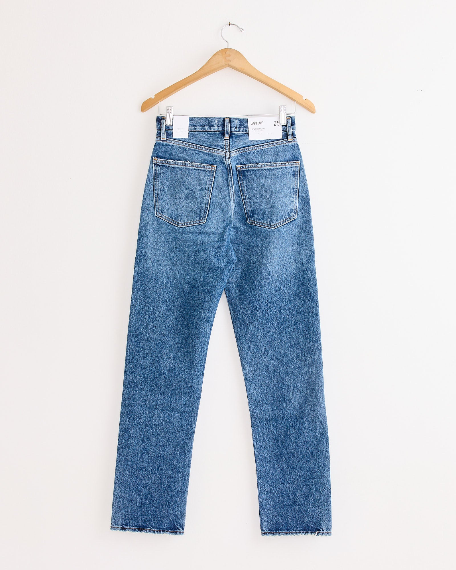 The AGOLDE 90's Pinch Waist Jean in Essence, crafted from blue regenerative cotton denim, hangs on a wooden hanger against a white background, shown from the back with two rear pockets and a straight-leg cut.