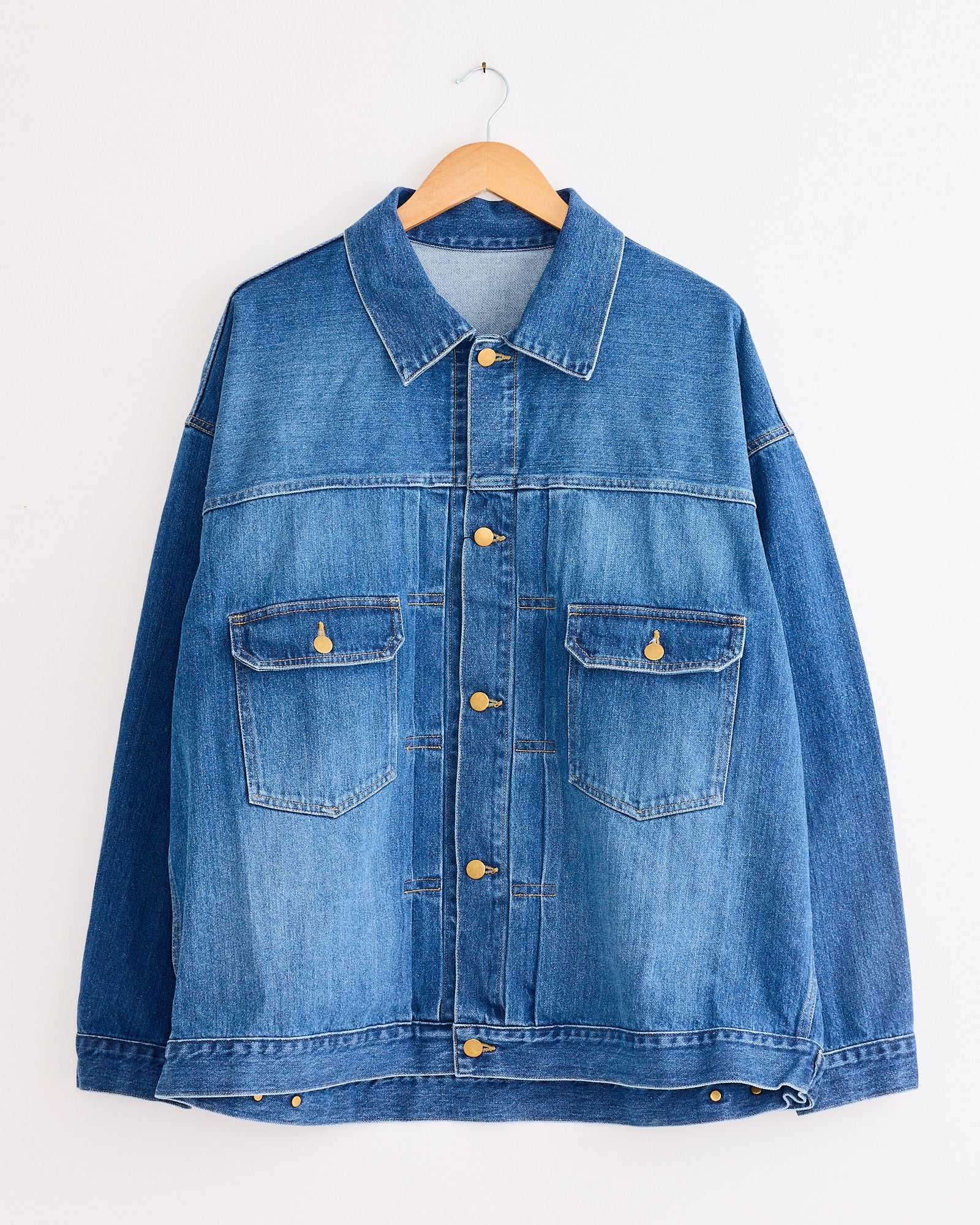 The Sillage Trucker Jacket in Blue Denim Damaged, featuring gold buttons and two front pockets, hangs on a wooden hanger against a white wall.