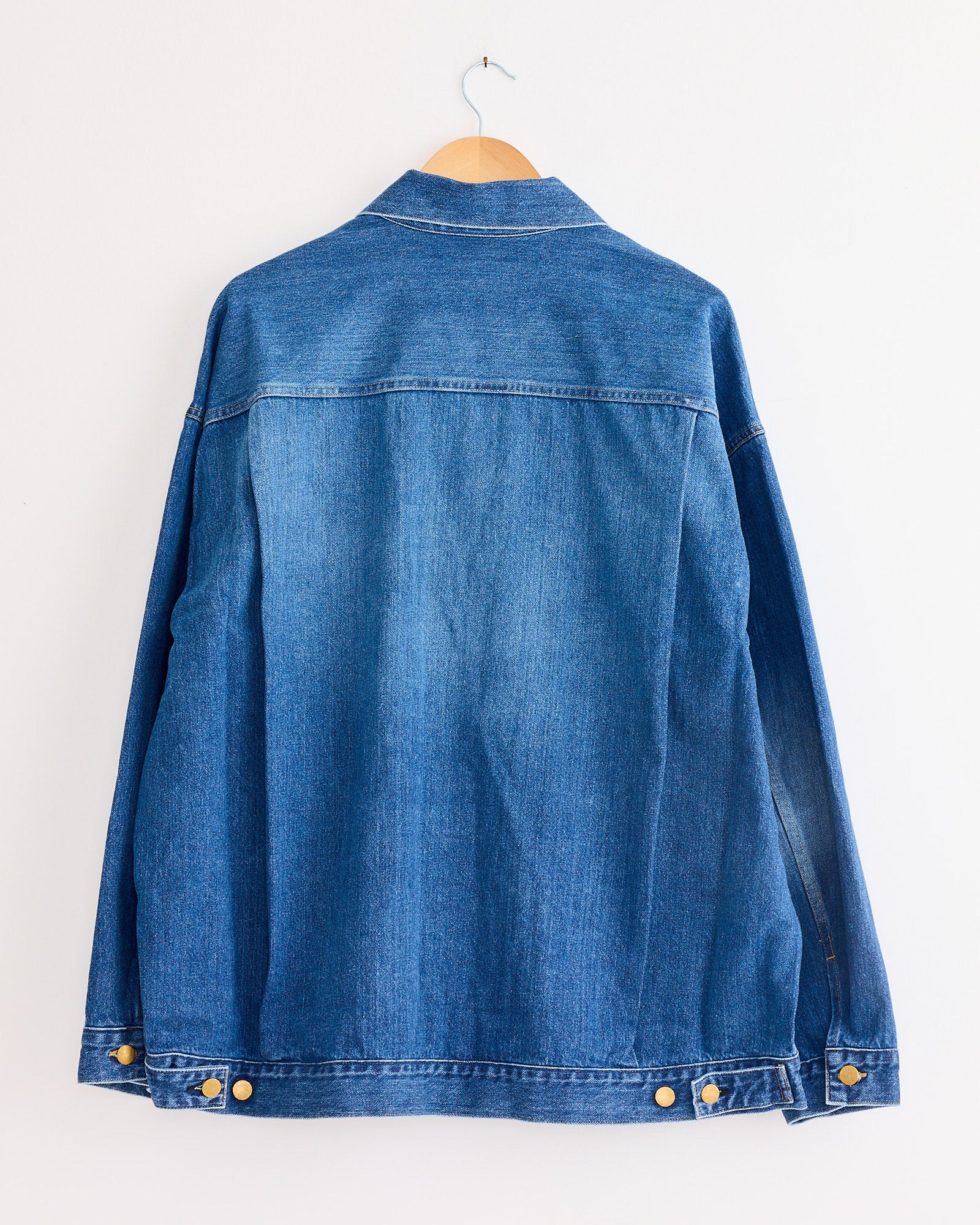 The Sillage Trucker Jacket in Blue Denim Damaged, made in Japan, hangs on a wooden hanger against a white wall. Displayed from the back, it has long sleeves with gold buttons at the cuffs and waistband.