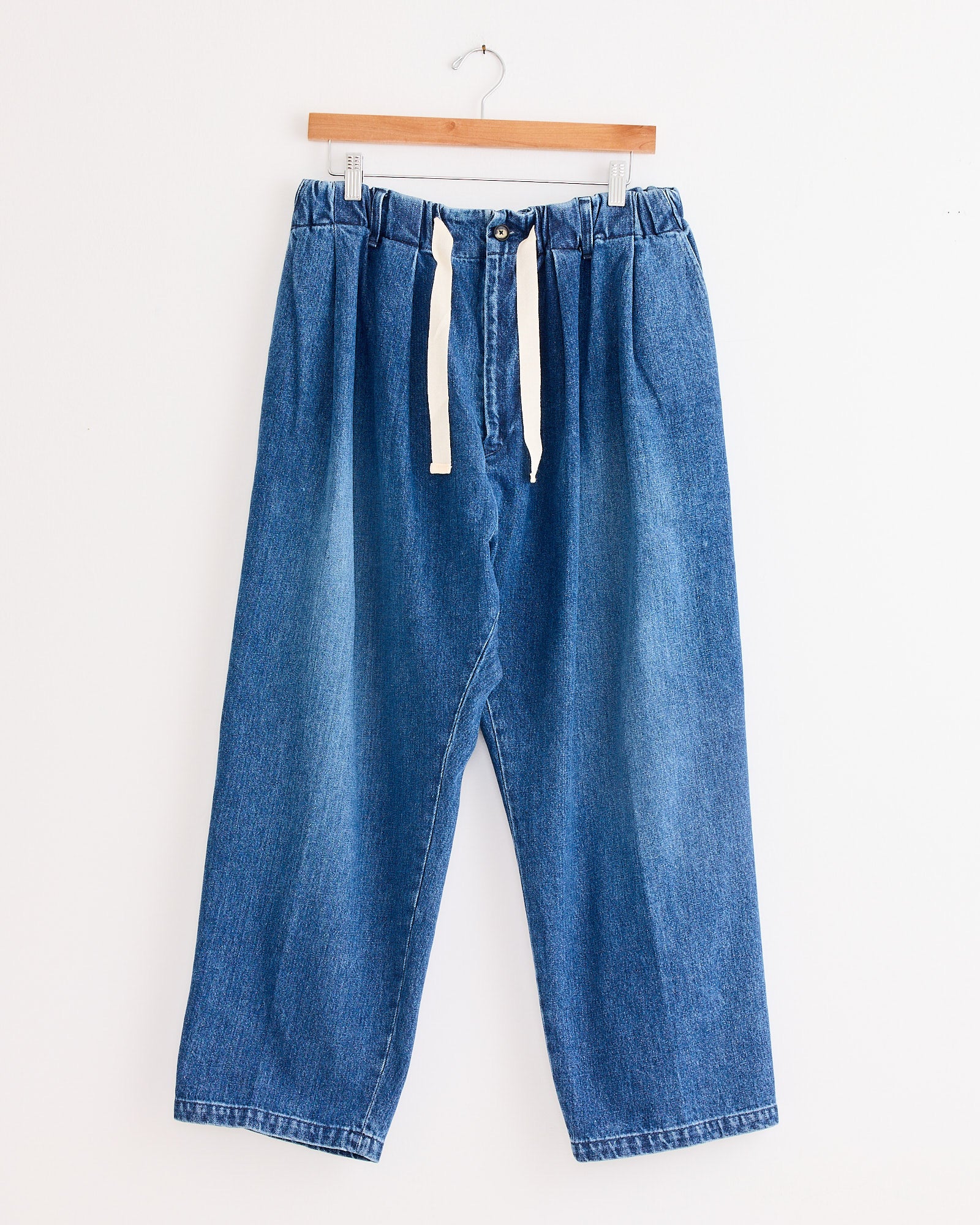 The Sillage Baggy Trouser in Blue Denim Damaged features a relaxed, loose fit with an elastic waistband and white drawstring, displayed on a wooden hanger against a plain white background.
