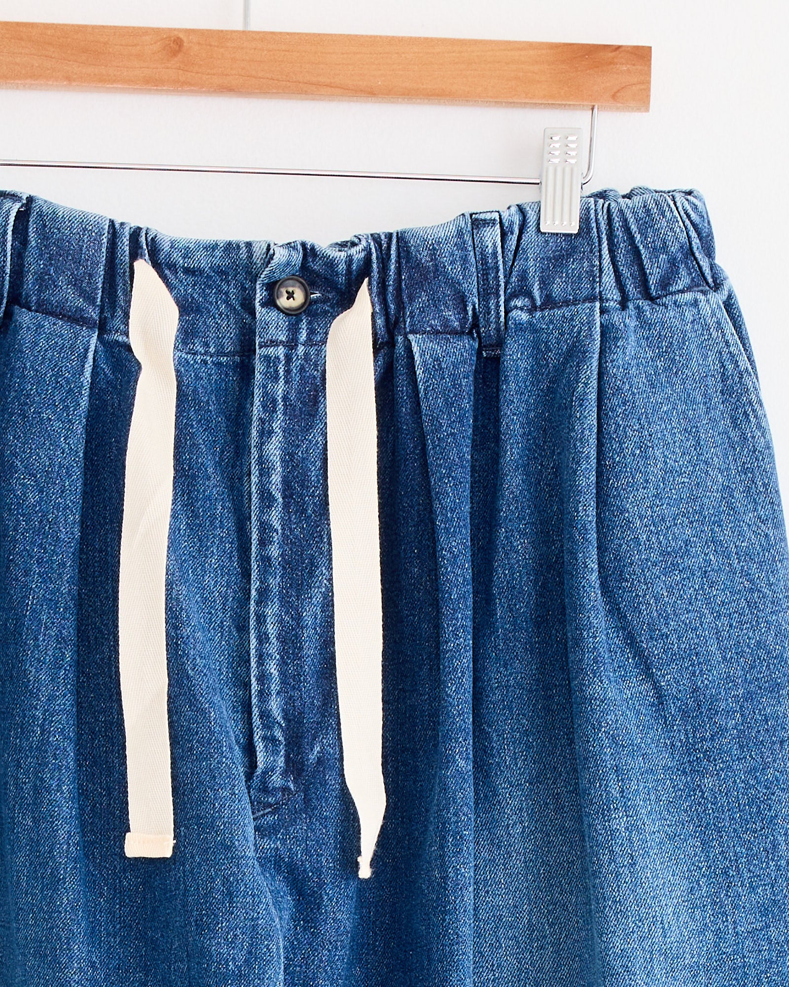 A close-up of Sillage’s Baggy Trouser in Blue Denim Damaged, featuring an elastic waistband, front button, and white drawstrings, hanging on a wooden hanger.