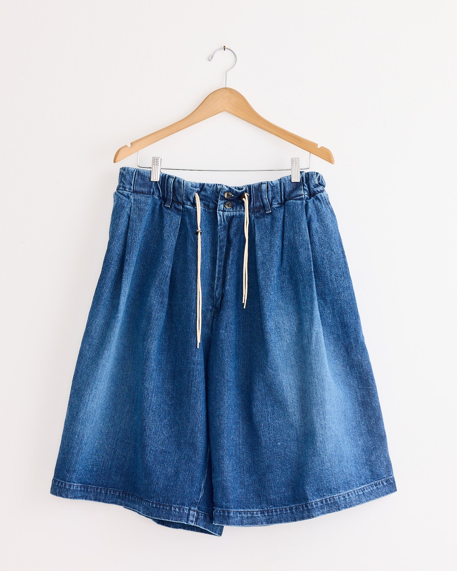 The Sillage Circular Short Pant Blue in Denim Damaged features an elastic waist, white drawstring, and wide legs—expertly made in Japan and displayed on a wooden hanger against a plain white wall.