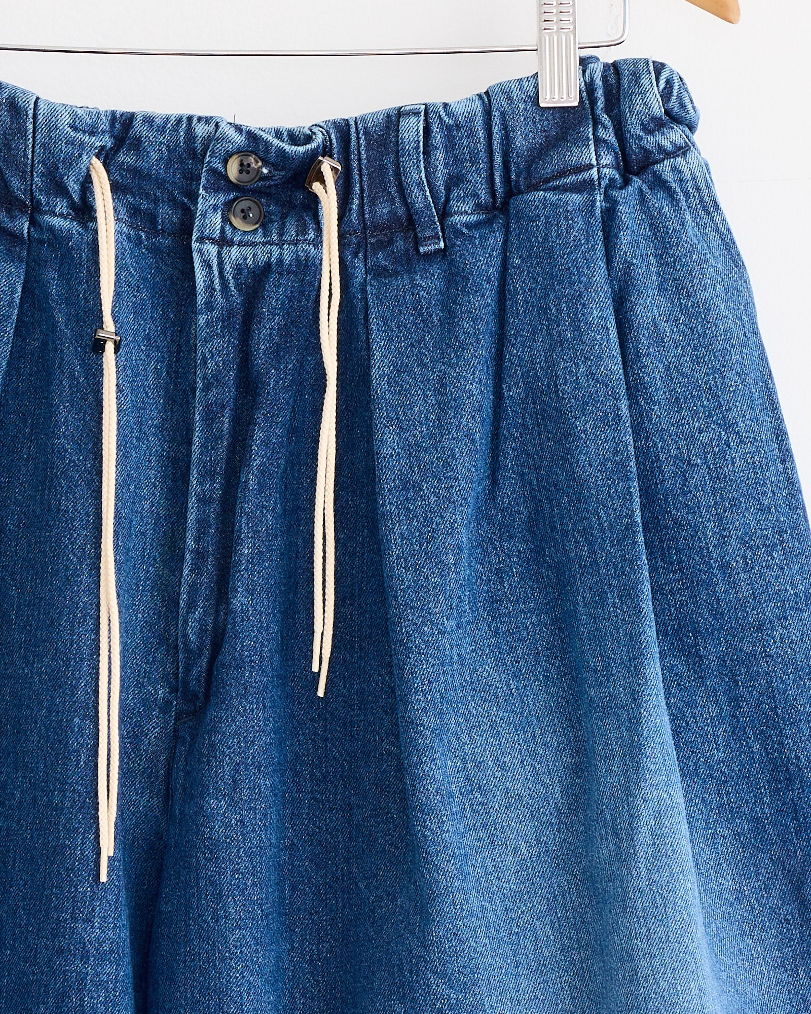 The Sillage Circular Short Pant Blue in Denim Damaged features an elastic waistband, two buttons, belt loops, and beige drawstrings. Made in Japan, it's shown hanging on a hanger against a white background.