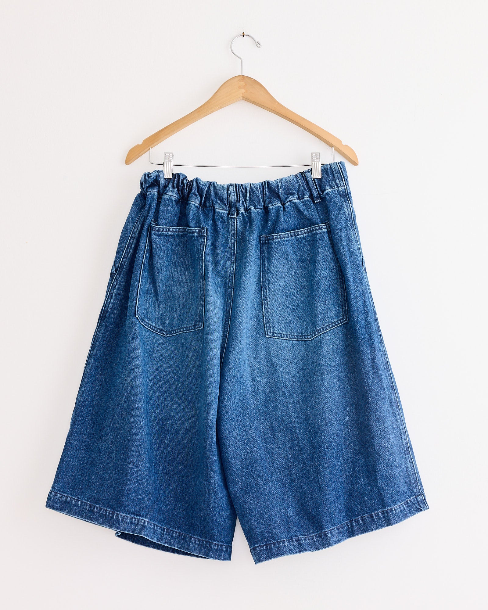 The Sillage Circular Short Pant Blue in Denim Damaged, featuring a wide-leg fit, elastic waistband, and two back pockets, hangs on a wooden hanger against a white wall.