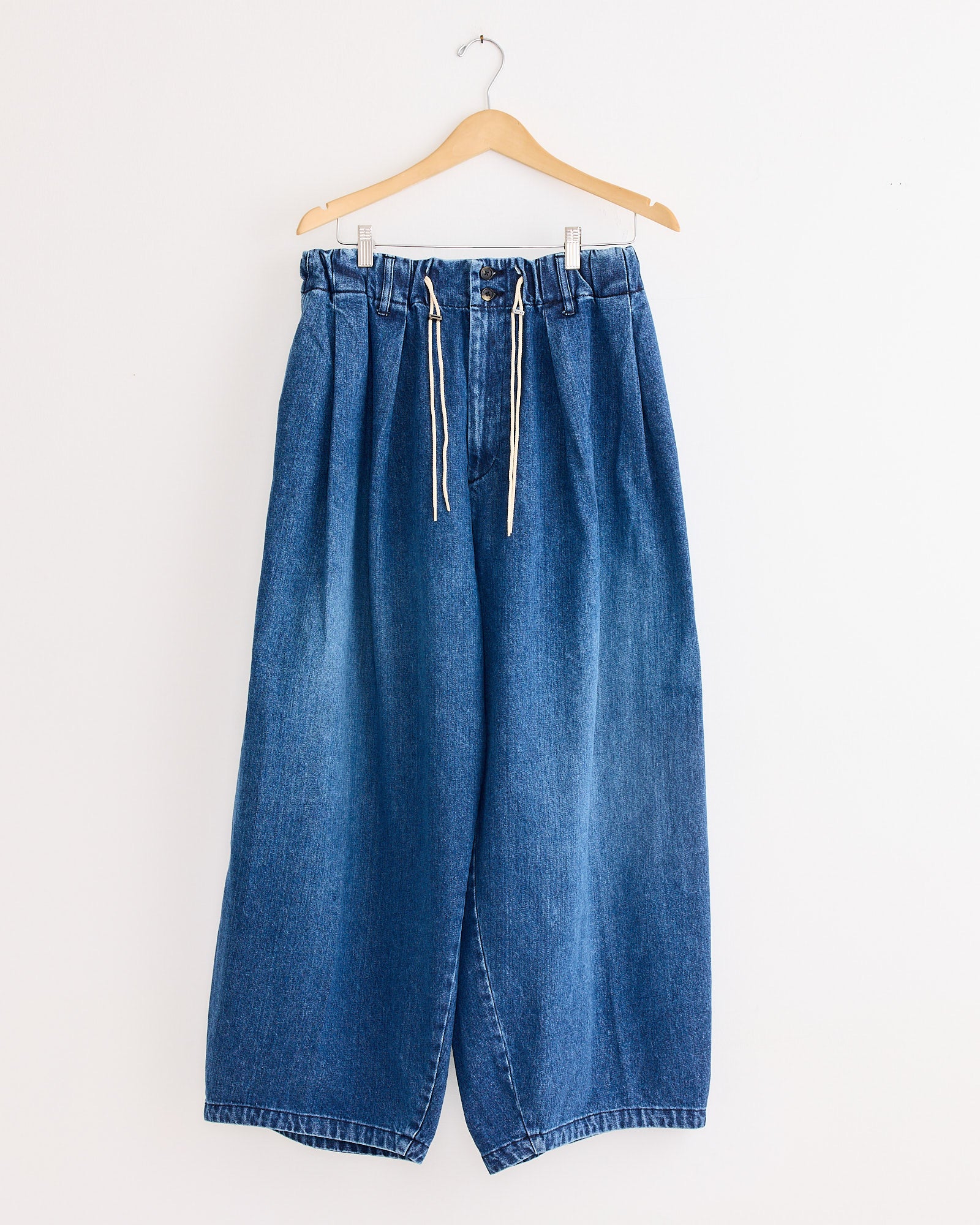 The Sillage Circular Pant Blue in Denim Damaged features a wide-leg fit, elastic waistband, and white drawstrings, shown hanging on a wooden hanger against a plain white wall.