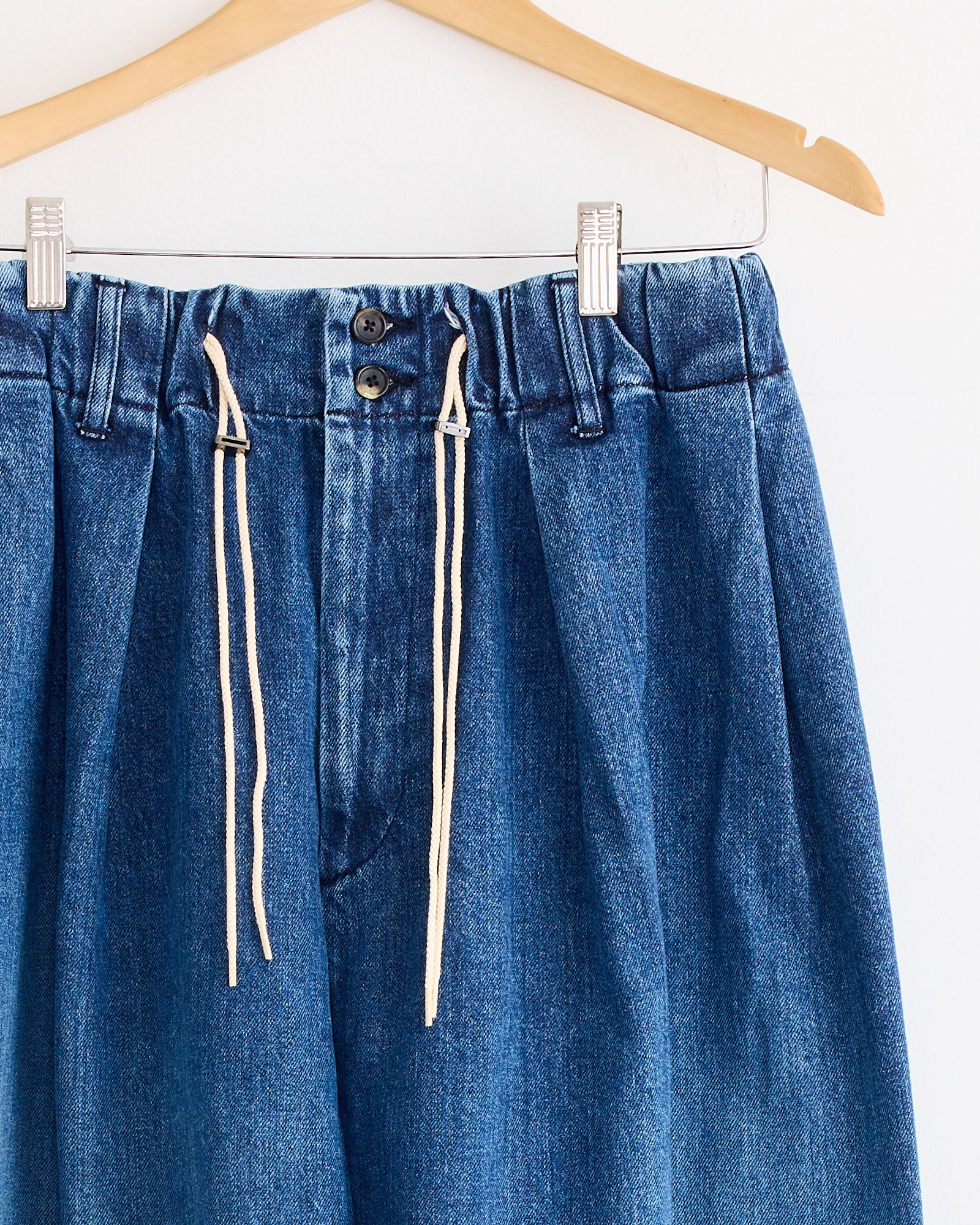 The Sillage Circular Pant Blue in Denim Damaged features an elastic waistband, drawstring ties, and two buttons, displayed on a wooden hanger against a white background.