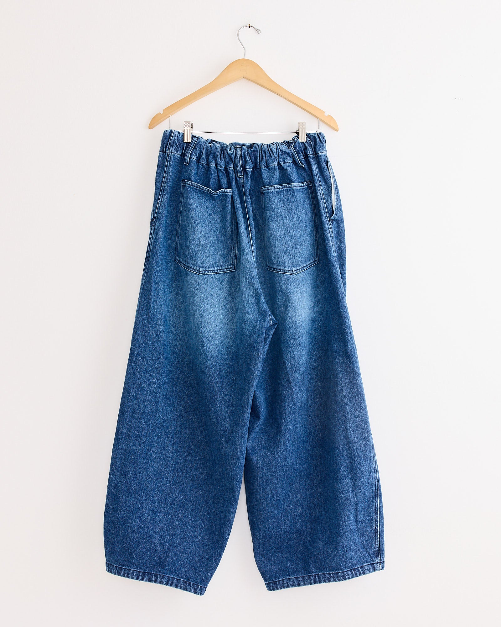 The Sillage Circular Pant Blue in Denim Damaged features wide legs, an elastic waistband, subtle distressed details, and two back pockets, displayed on a light wooden hanger against a plain white wall.