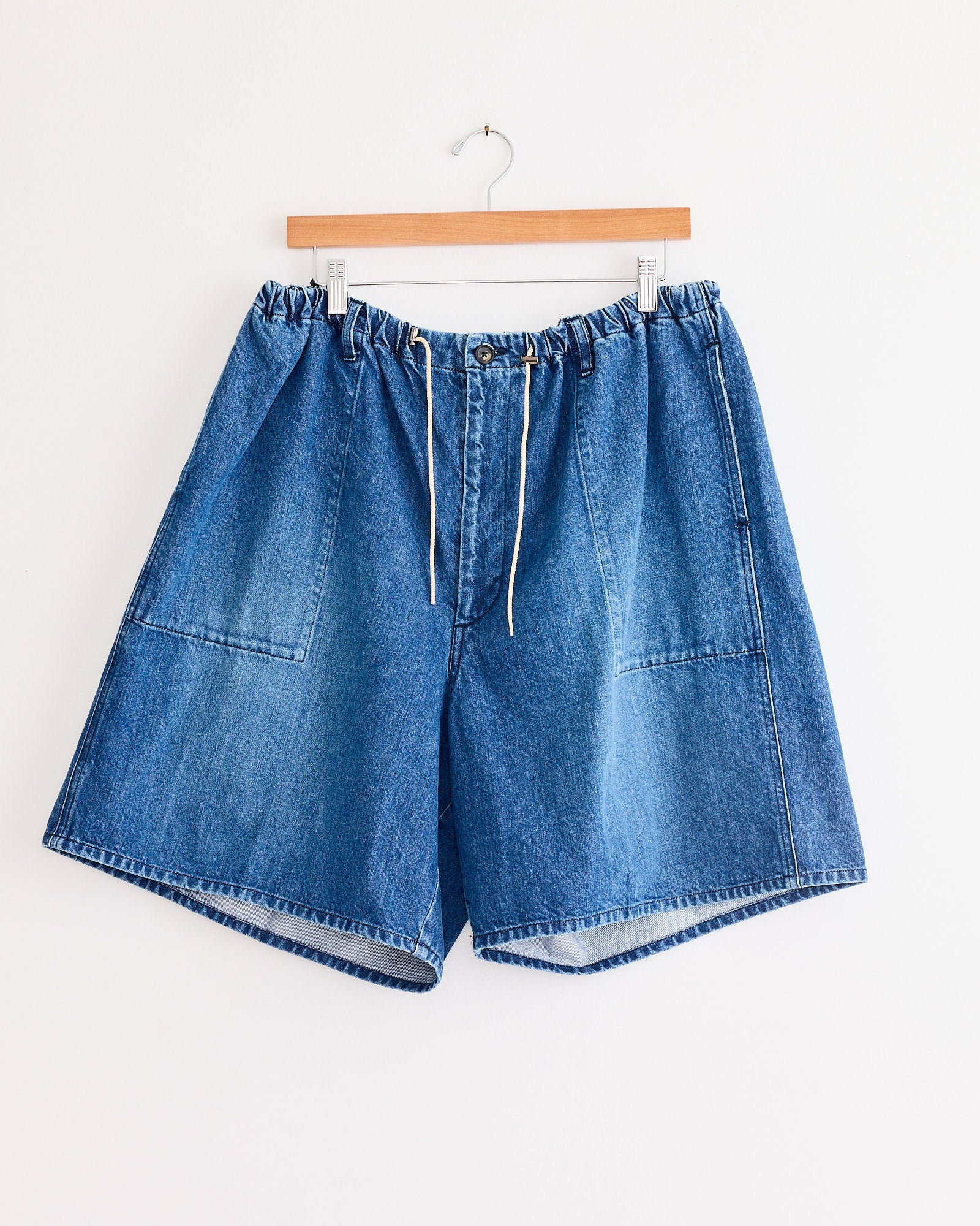 The Sillage Circular Fatigue Short in Blue Denim Damaged, made from organic cotton denim with an elastic waistband, drawstring, zipper, and large front pockets, hangs on a wooden hanger against a white wall.