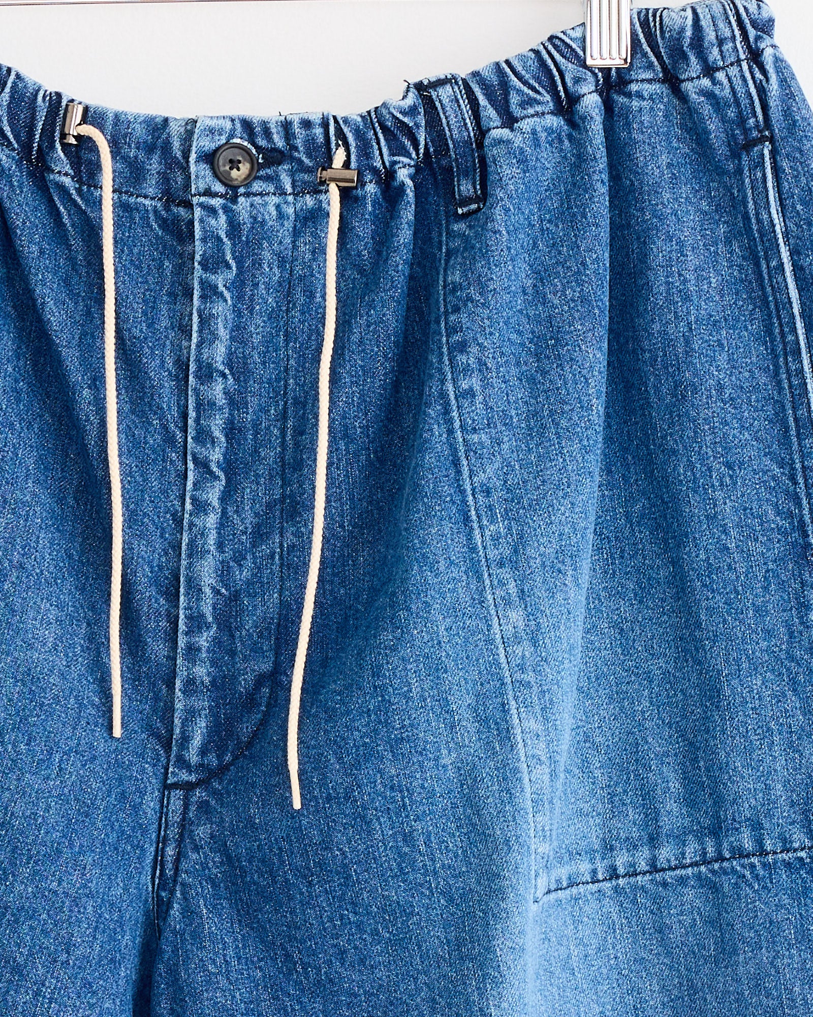 Close-up of Sillage Circular Fatigue Short in Blue Denim Damaged, made in Japan with an elastic waistband, white drawstring, button closure, and crafted from organic cotton denim with visible stitching details on the front and pockets.